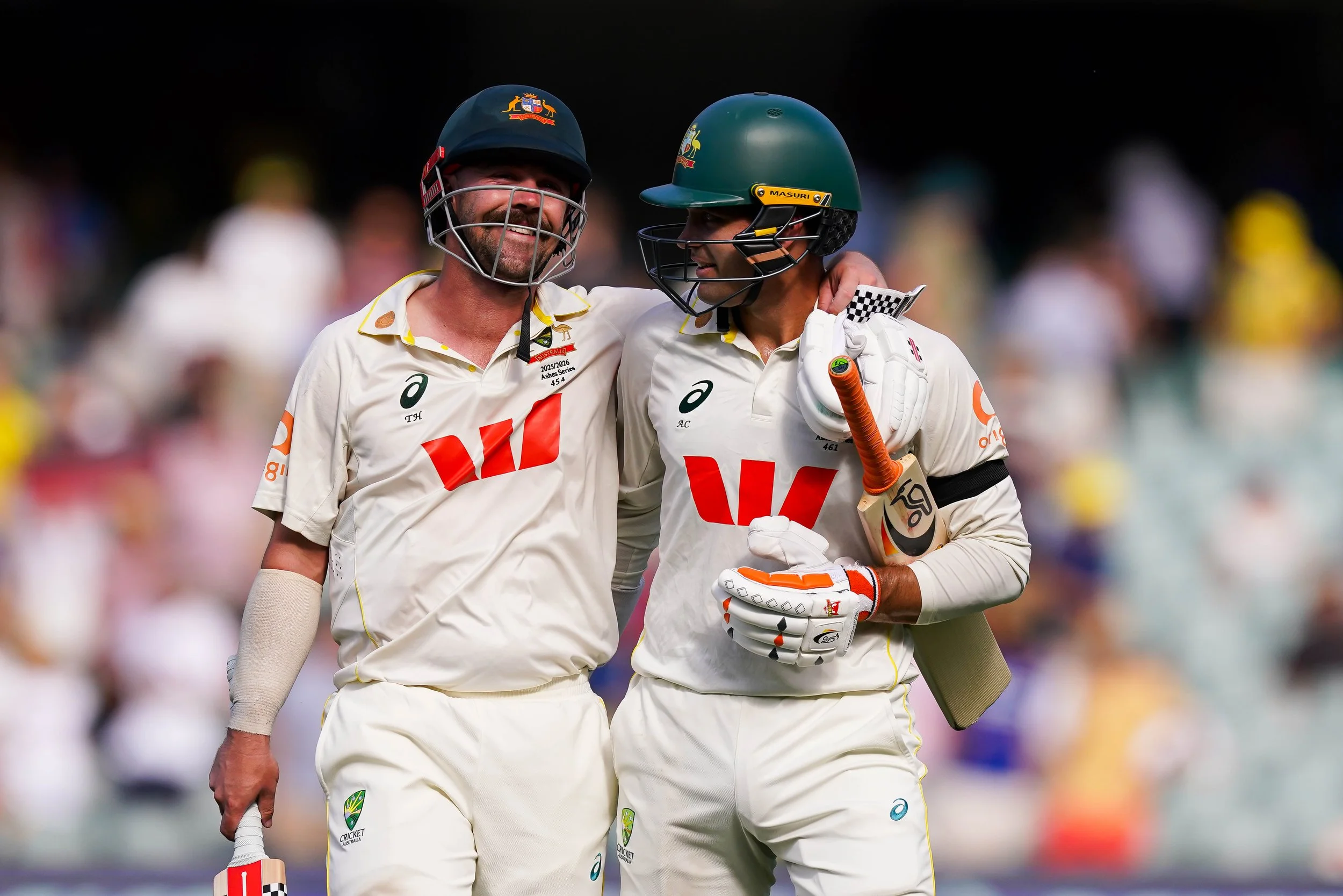 Two cricket players in white uniforms smiling and celebrating on the field. They are wearing green and dark blue helmets and gloves, with one holding a cricket bat. The background shows blurred spectators.