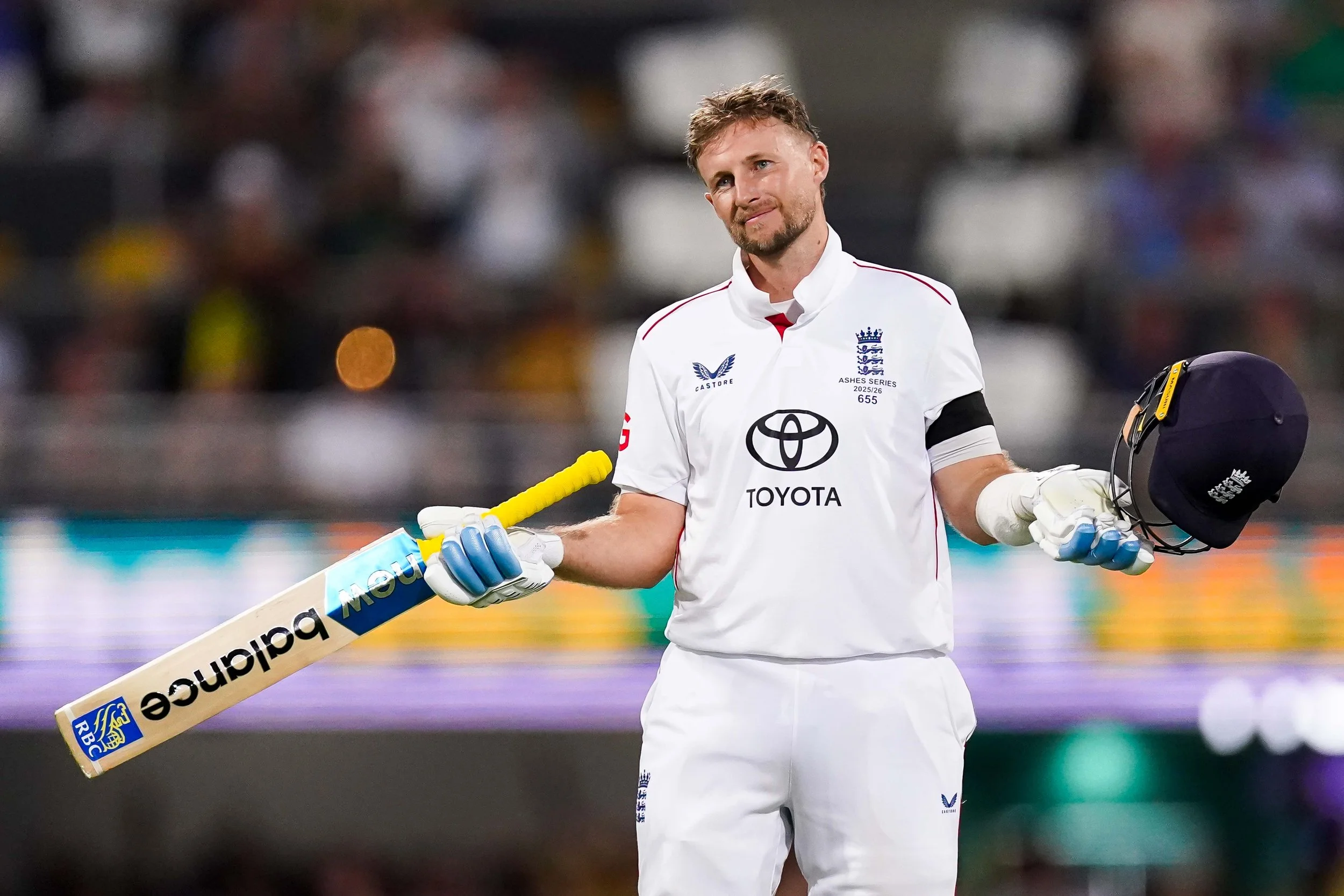 Joe Root of England in white uniform holding a cricket bat and helmet on a cricket field, celebrates his first century in Australia at The Gabba, Brisbane.
