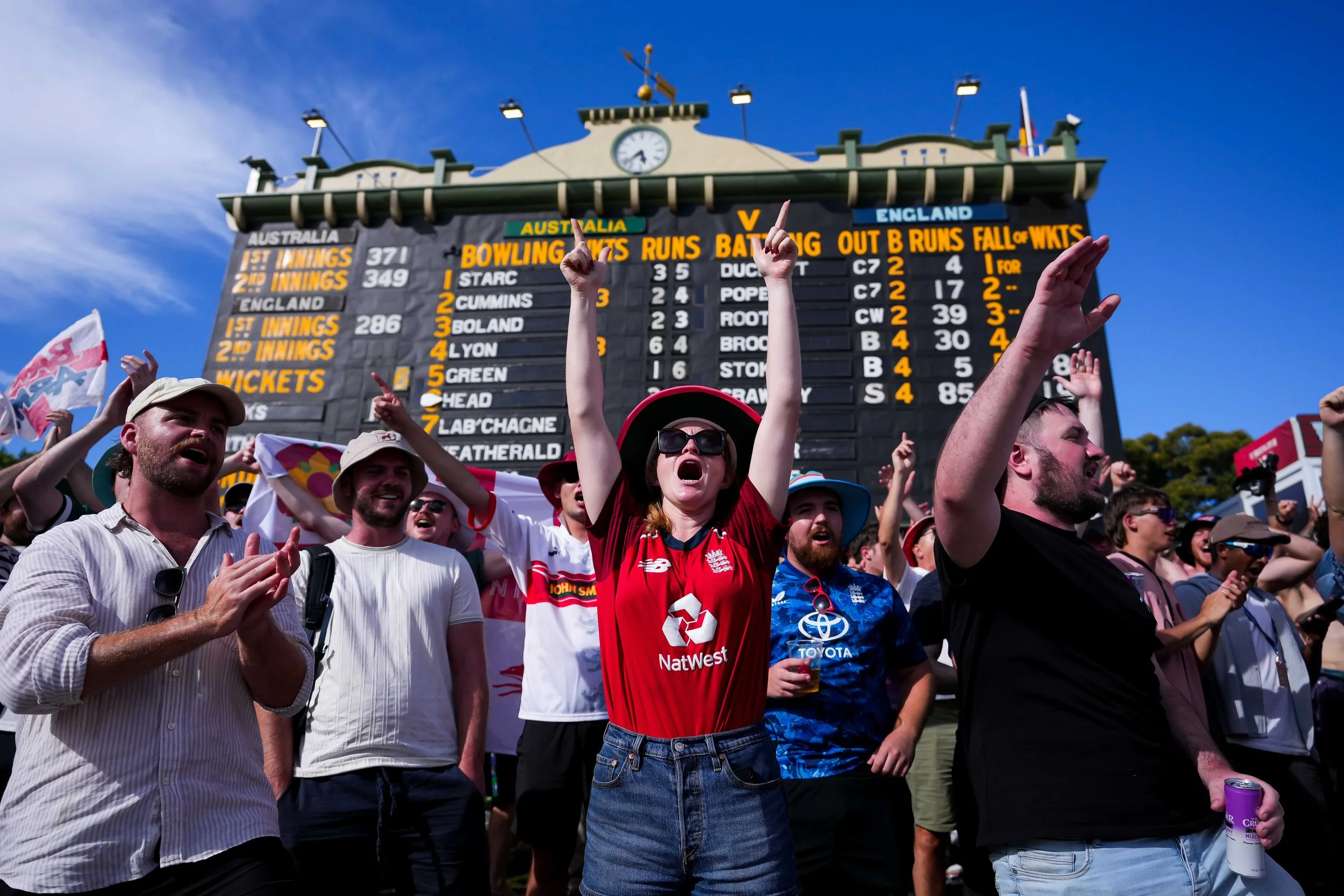 Group of cricket fans cheering at The Adelaide Oval during daytime with scoreboard in the background.