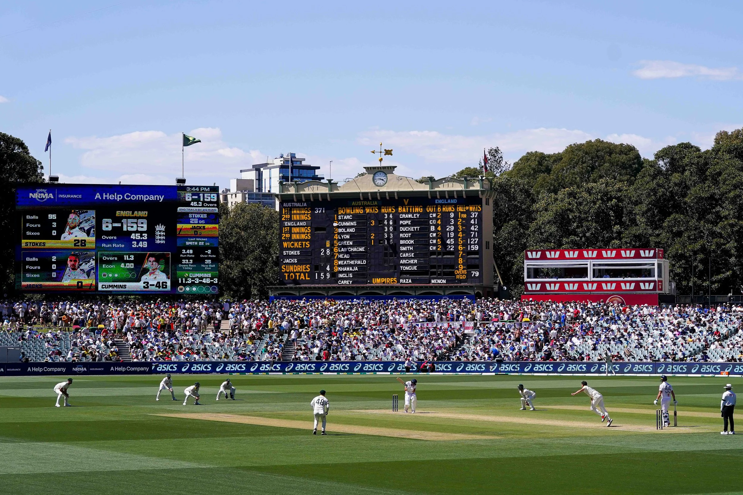 Cricket match at a stadium with players on the field and a crowd in the stands, large scoreboard showing scores and statistics, blue sky with some clouds.