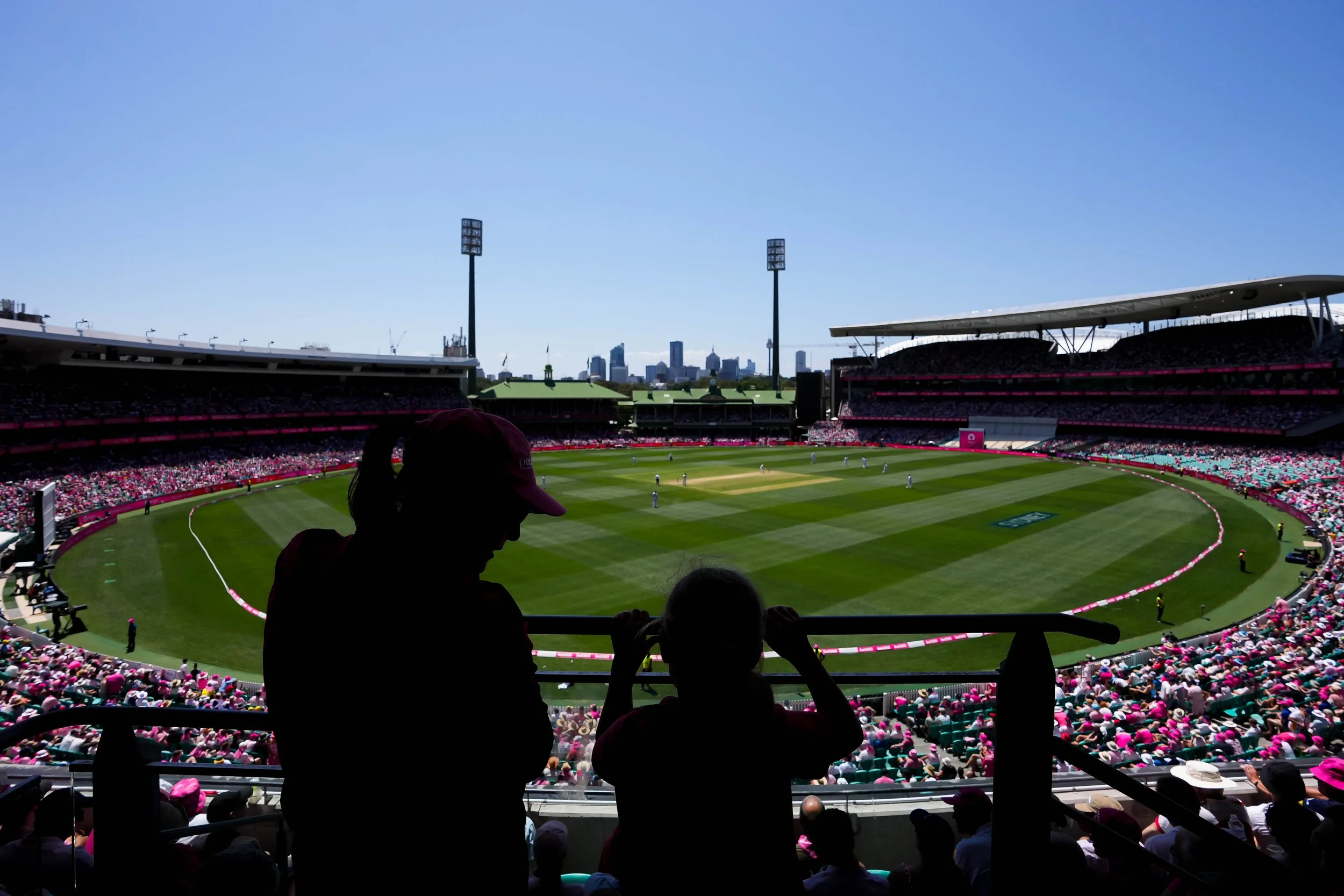 Silhouetted persons watching a baseball game from stadium seats with a city skyline in the background.