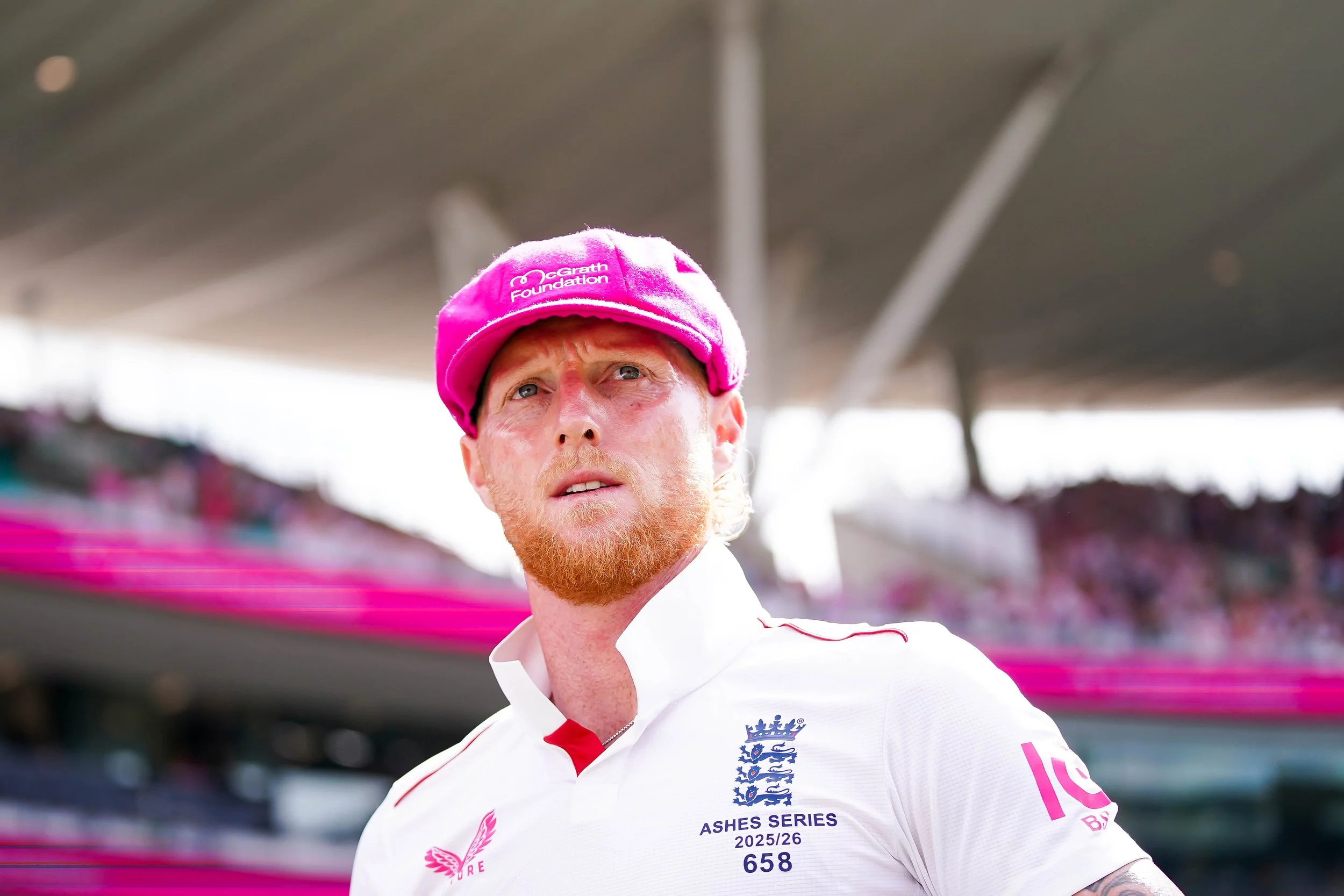 A male cricket player wearing a pink cap and white jersey with blue and pink logos, standing on a cricket field under a stadium roof.