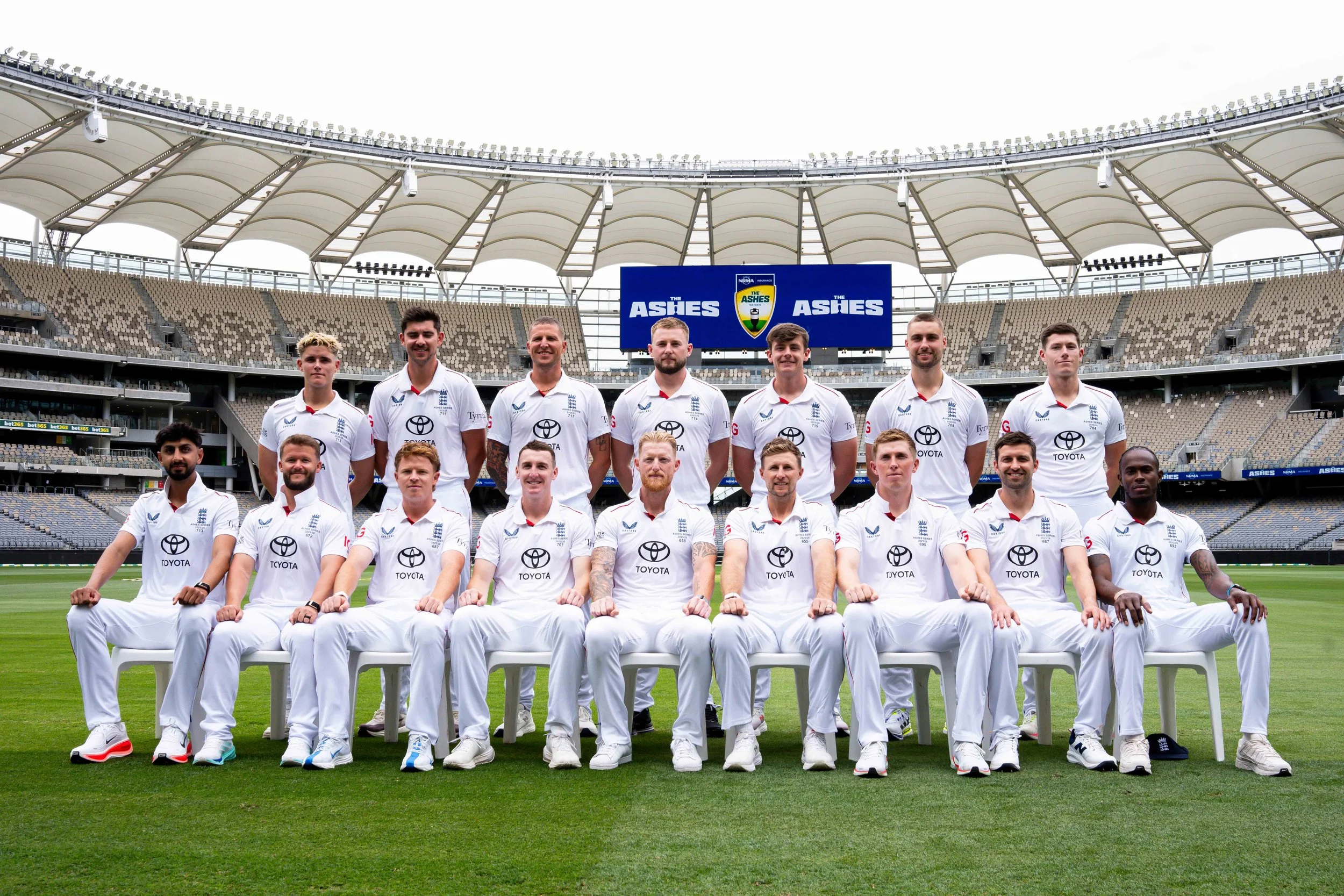 A cricket team poses for a photo in a stadium with a large digital screen behind them displaying 'The Ashes' logo, with eleven players, some sitting and some standing, dressed in white uniforms.