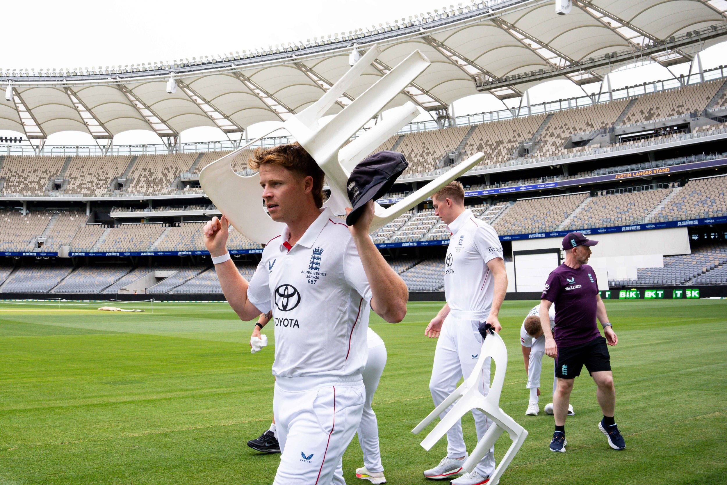 Cricketers in white uniforms walking on a cricket field, carrying white chairs, with a stadium in the background.