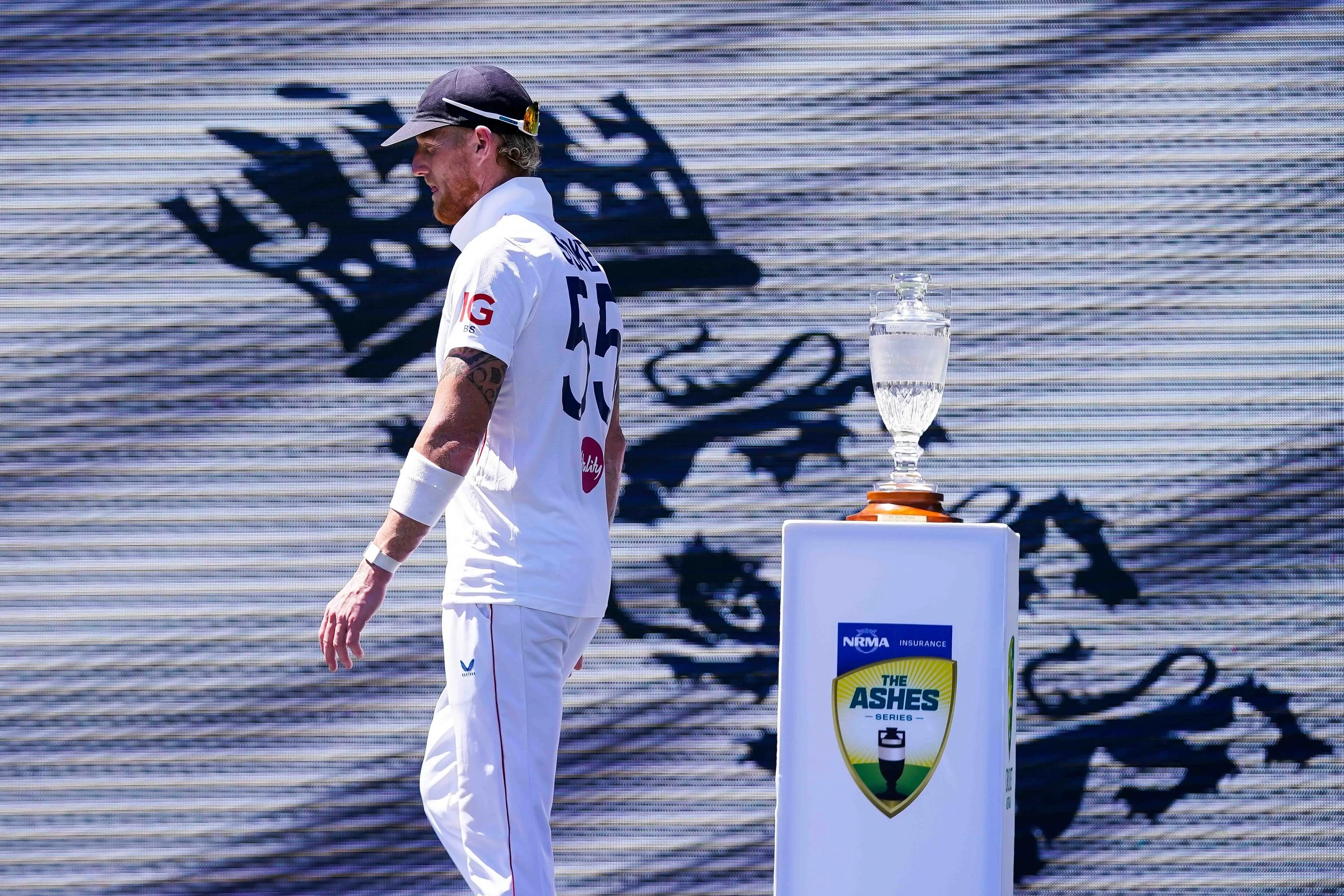 A man wearing a cricket uniform stands next to a trophy on a pedestal, with a large screen in the background displaying a cricket logo and the words 'The Ashes' along with sponsorship logos.