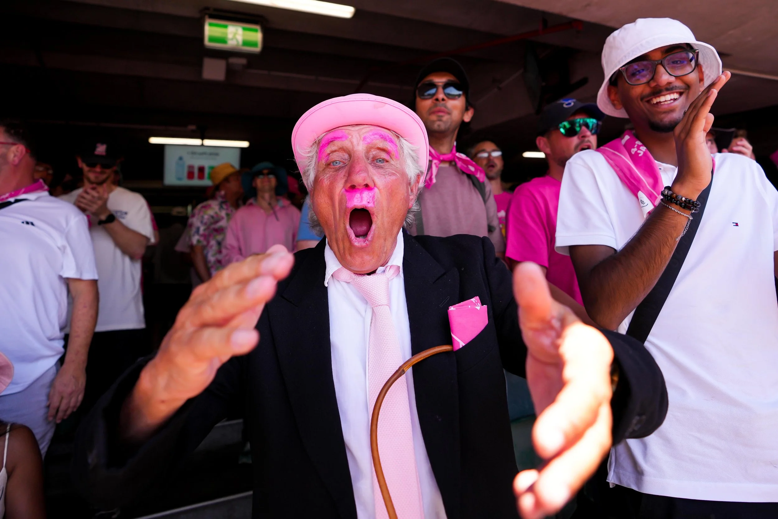 A group of people at an outdoor event wearing pink and white clothing, with some wearing hats and sunglasses. An elderly man with painted pink face and white hair, wearing a pink hat, is in the foreground with her mouth open and hands reaching out.
