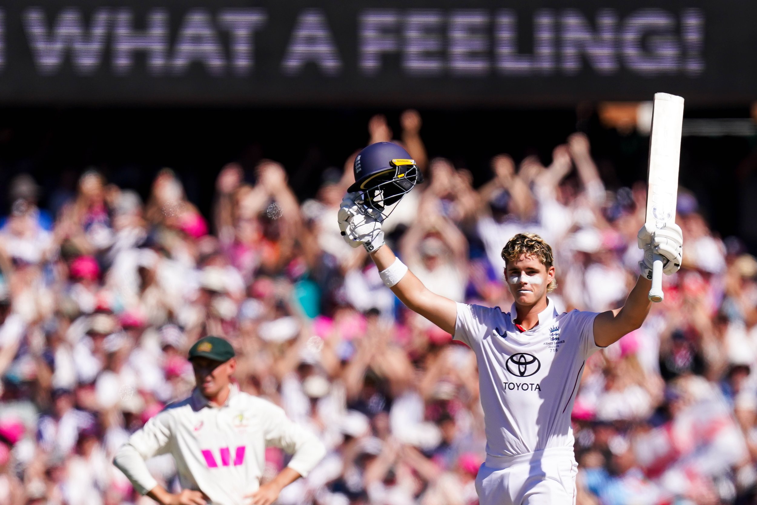 Cricketer celebrating with a raised helmet and bat during a cricket match with a crowd in the background.