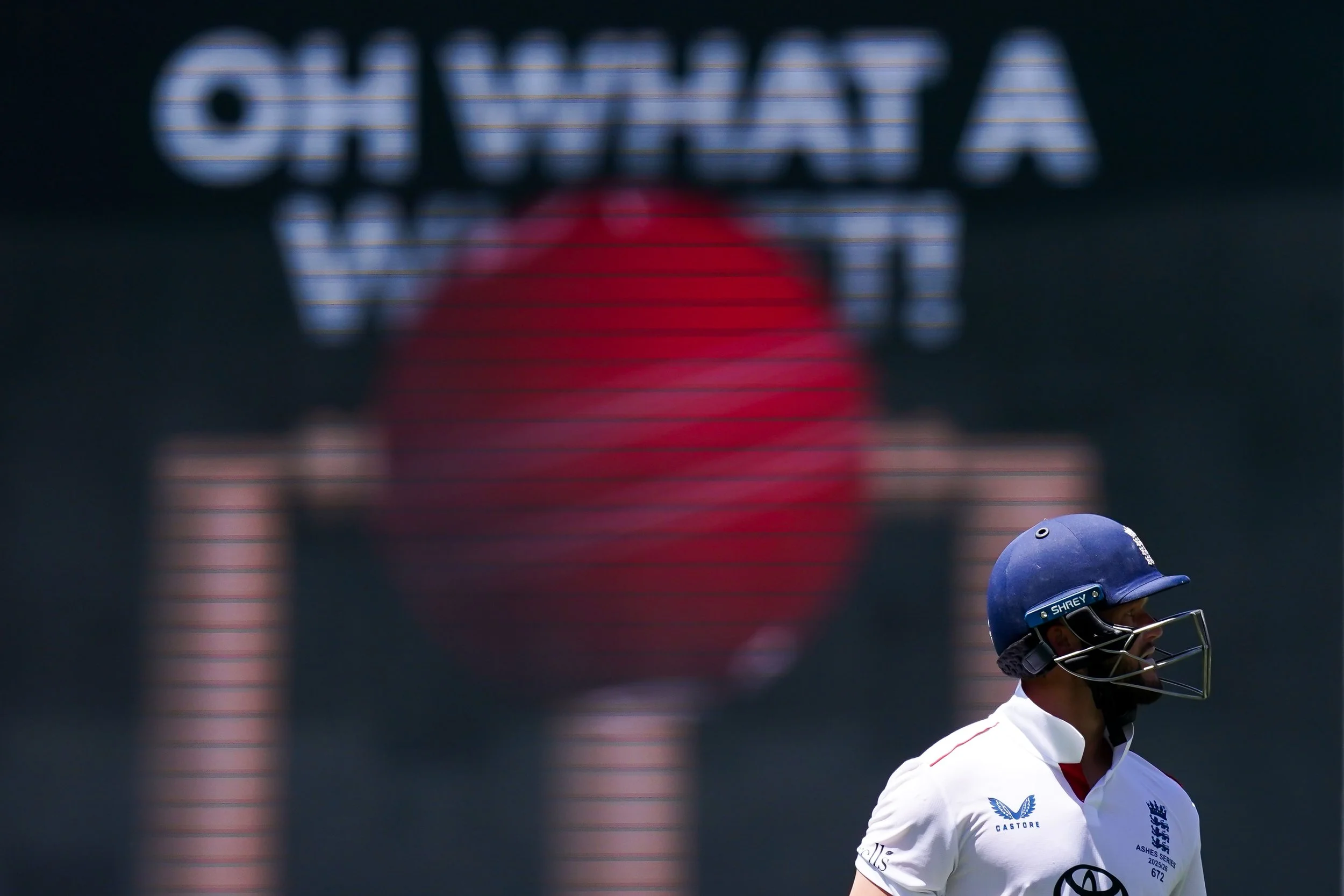 A cricket player in a white uniform and blue helmet walking on the field with a scoreboard in the background showing a large red ball and the name 'England'.
