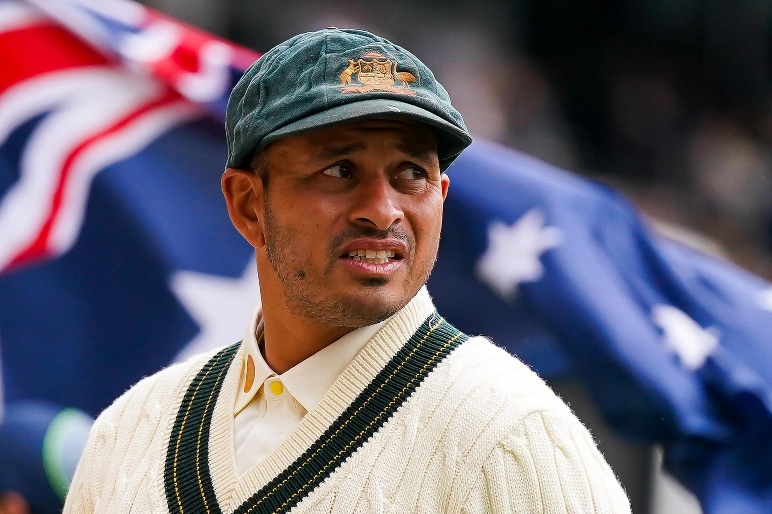 Usman Khwaja, wearing a baggy green cap and a cream-colored sweater, standing in front of an Australia flag.