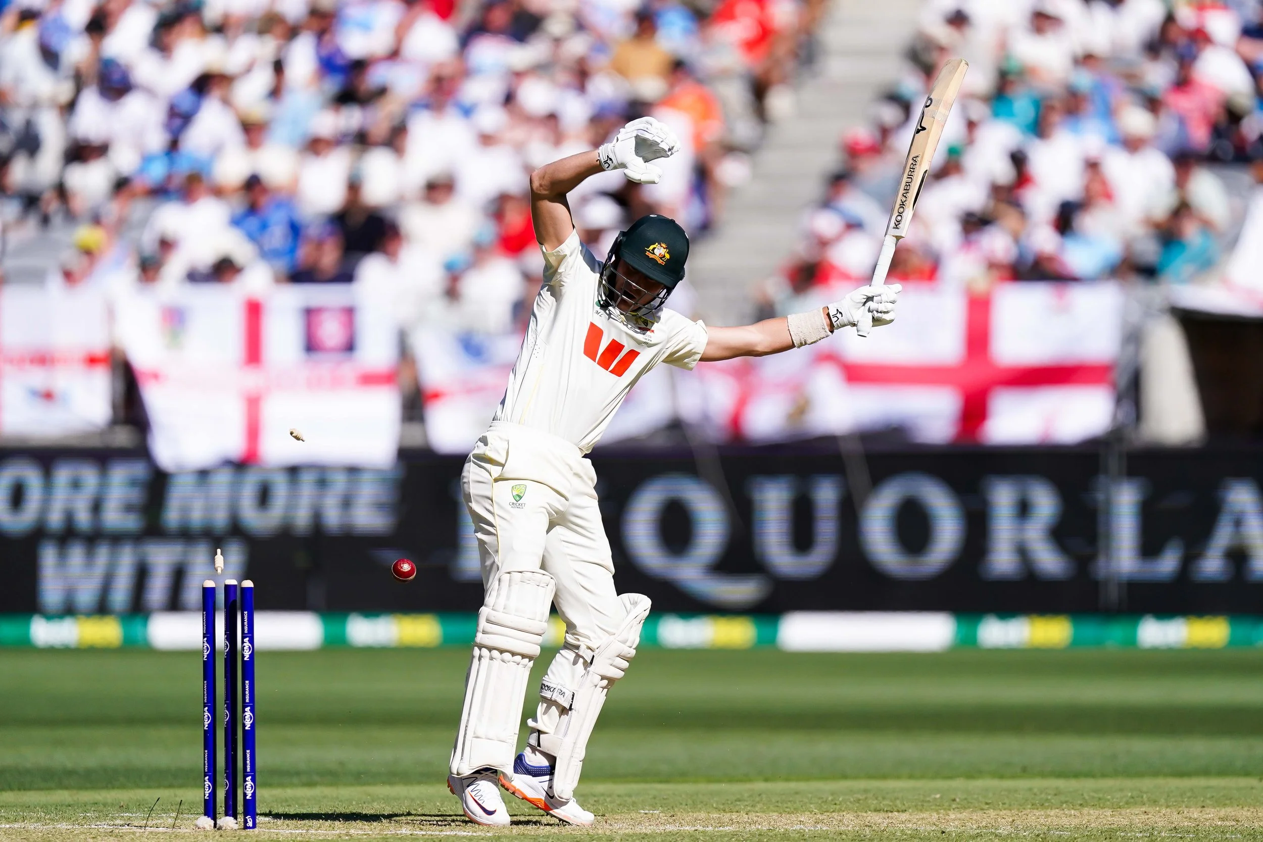 A cricket player in white uniform hitting a red cricket ball with a bat on a cricket pitch during a match with a crowd of spectators in the background.