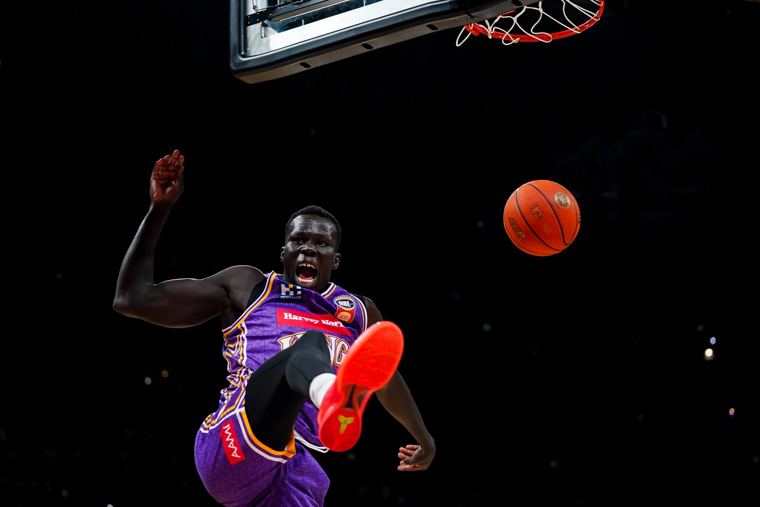 A basketball player in a purple uniform is mid-air, about to strike a loud defensive pose as a basketball bounces in the air towards the hoop, with a dark background.