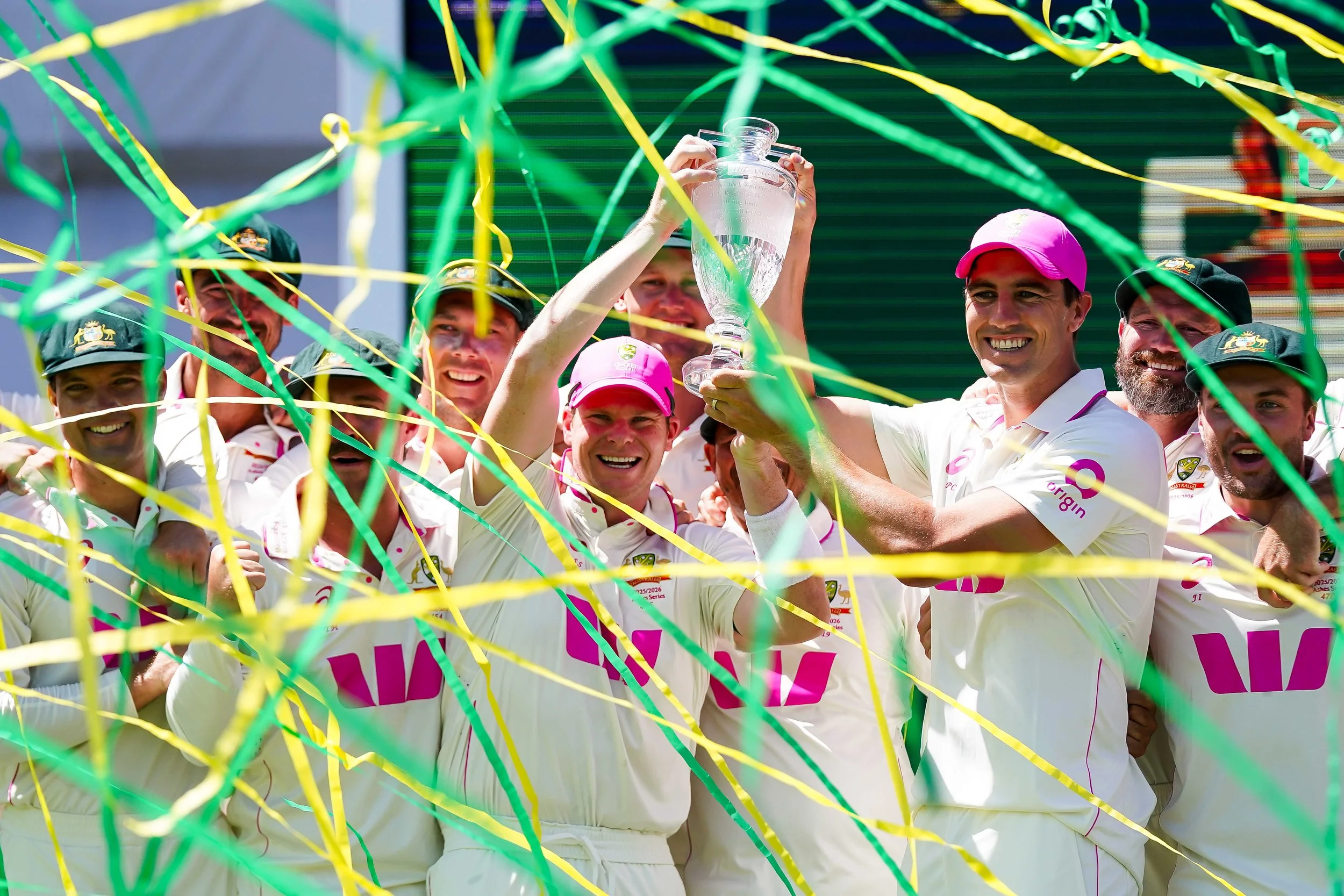 Australian cricket team celebrating after winning a match, with one player holding a trophy, decorated with green and yellow streamers and confetti.