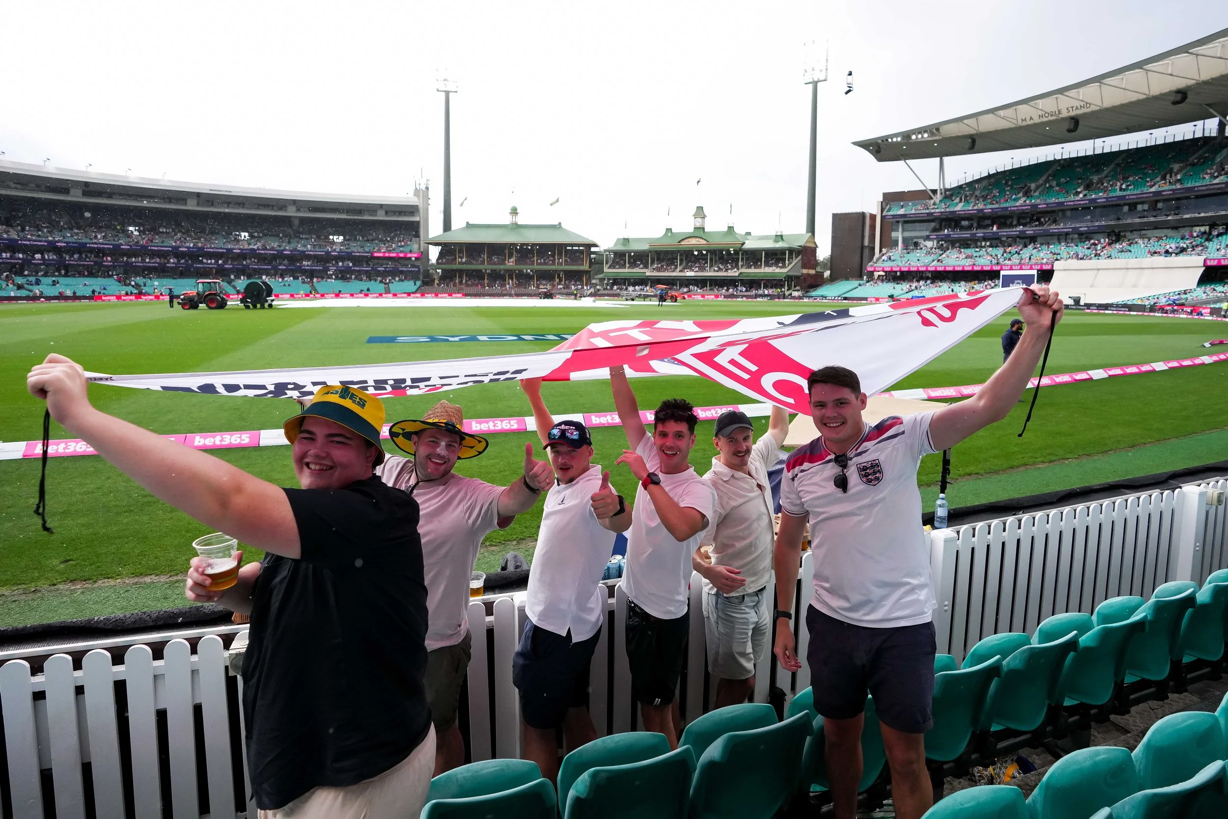 Group of six friends celebrating at a cricket stadium, holding a large England cricket team banner, with drinks in hand and smiling.