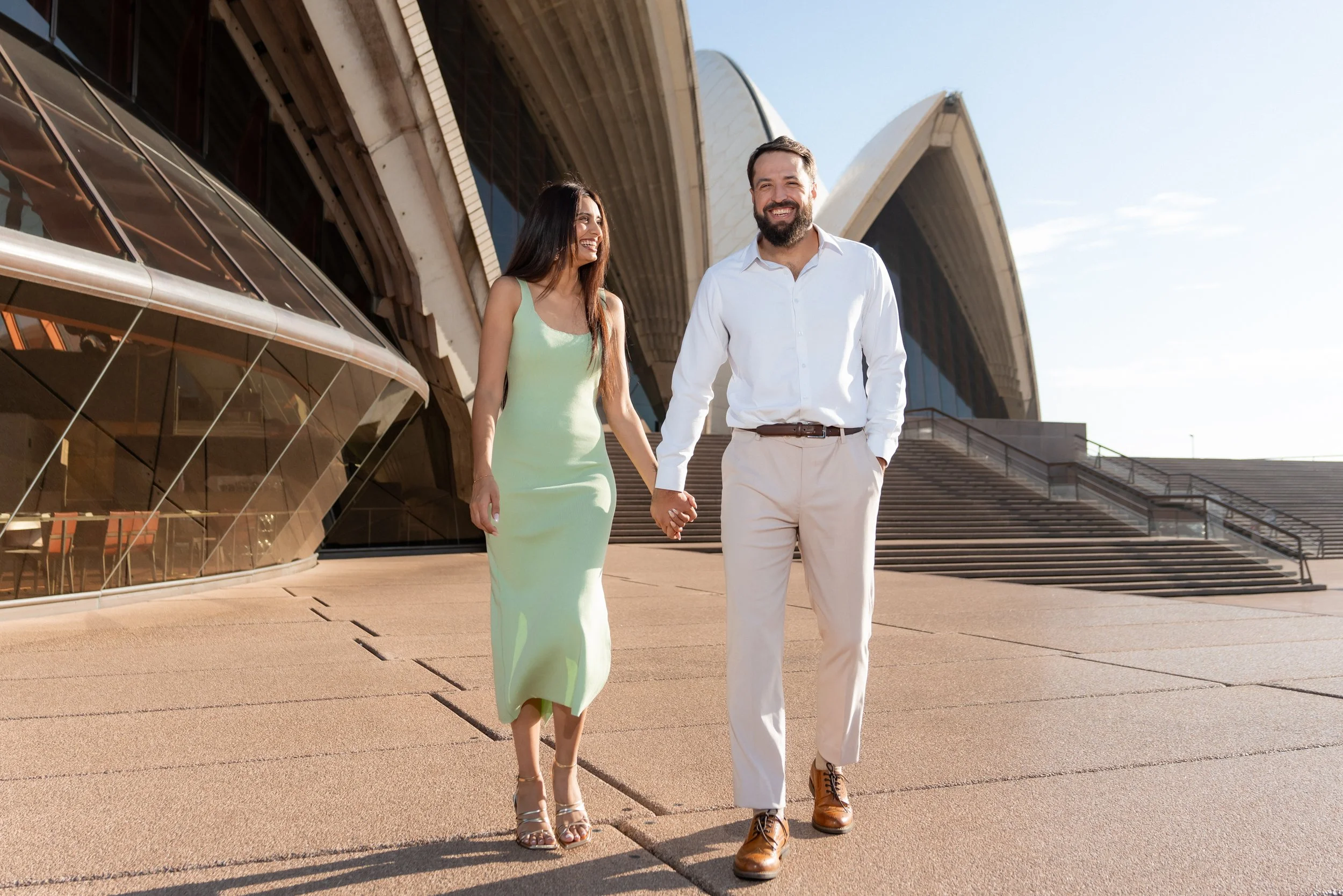 A couple walking hand in hand outside the Sydney Opera House, smiling.