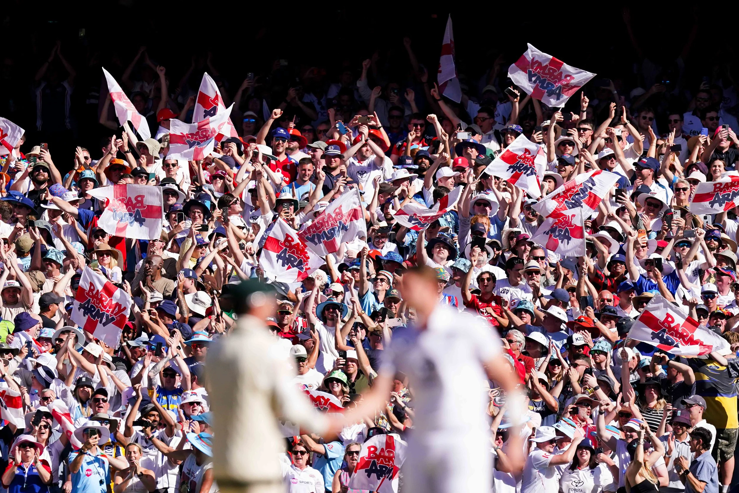 Crowd of race fans at a sporting event, holding flags and taking photos, with two blurred figures in the foreground.