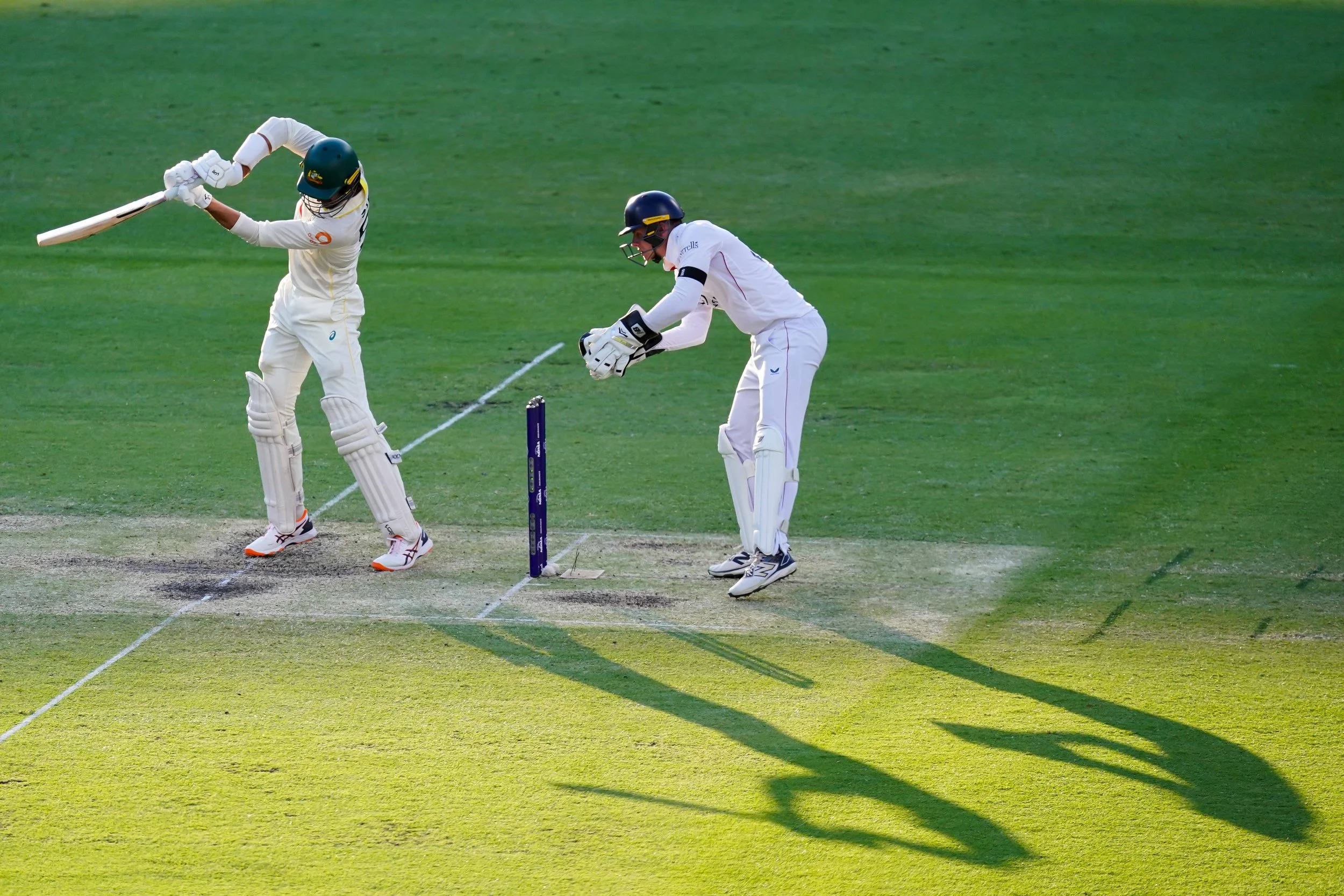 Cricket players in whites, one hitting the ball, another fielding or wicket-keeping on a grassy field.