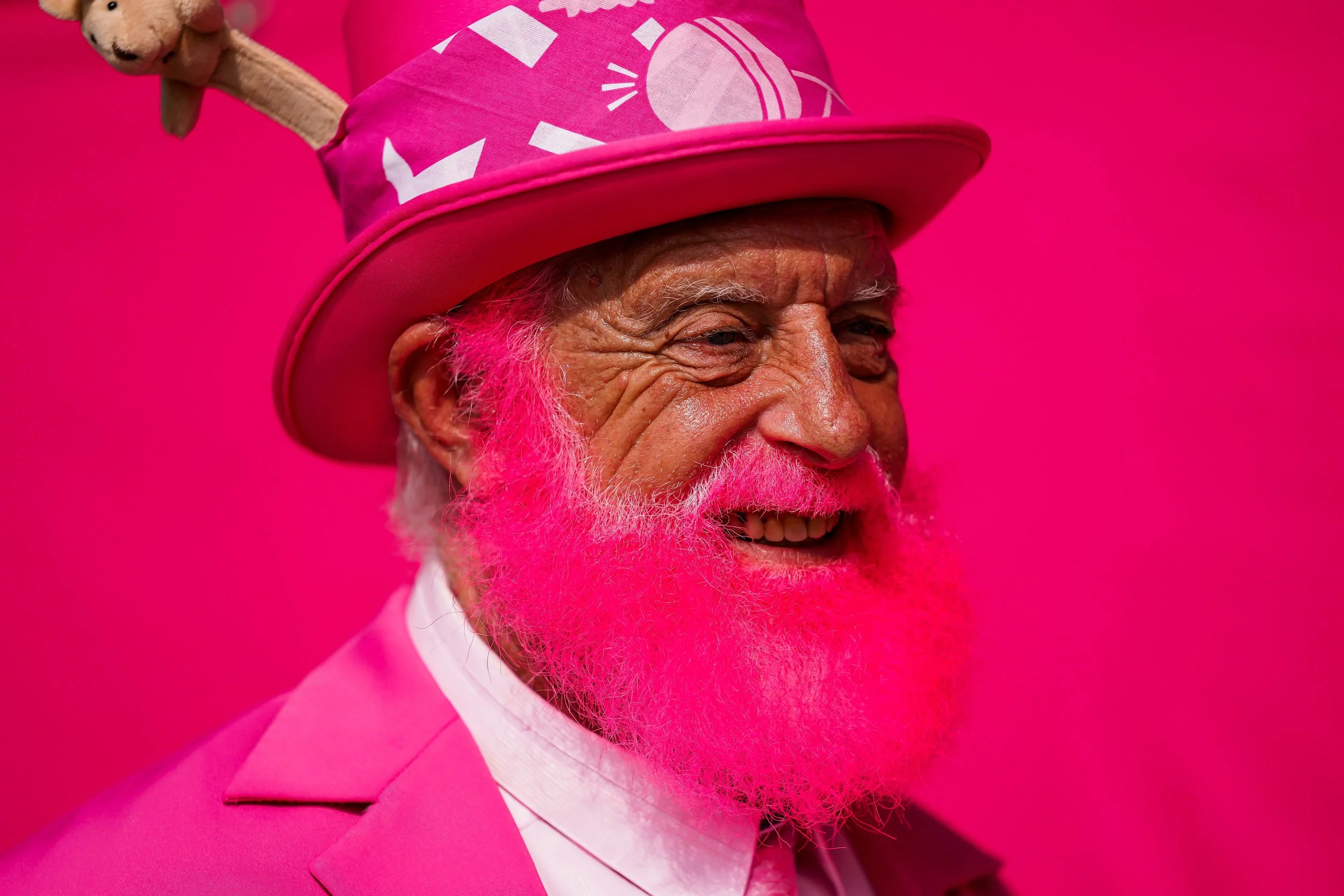 A smiling elderly man with a bright pink beard, pink hat, and pink suit against a pink background.