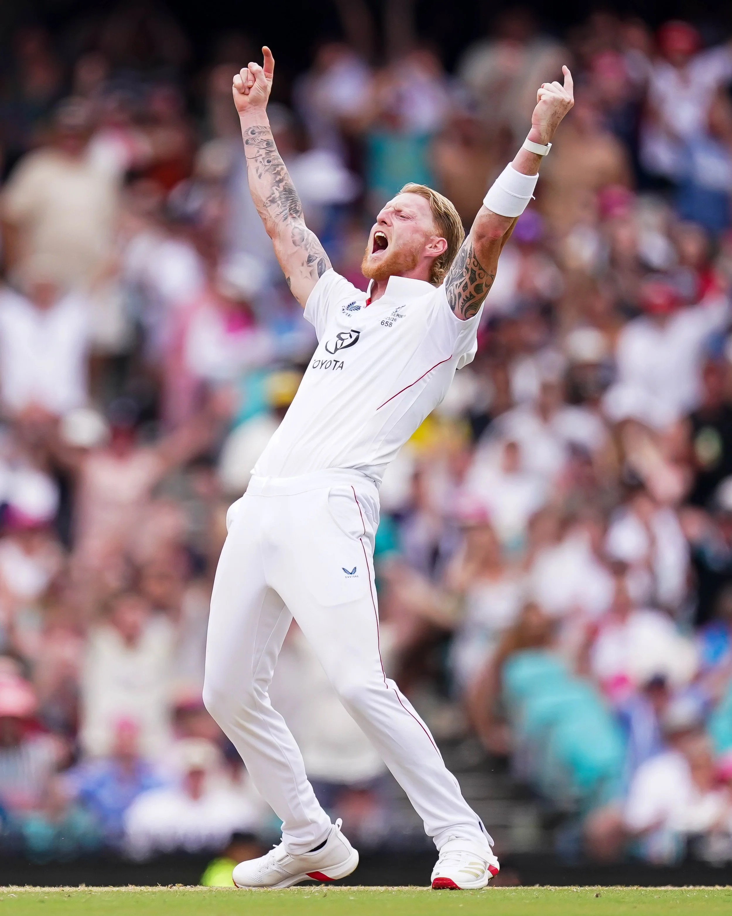 A cricket player wearing a white uniform with tattoos on his arms, celebrating with his arms raised in victory on a cricket field, with a crowd of spectators in the background.