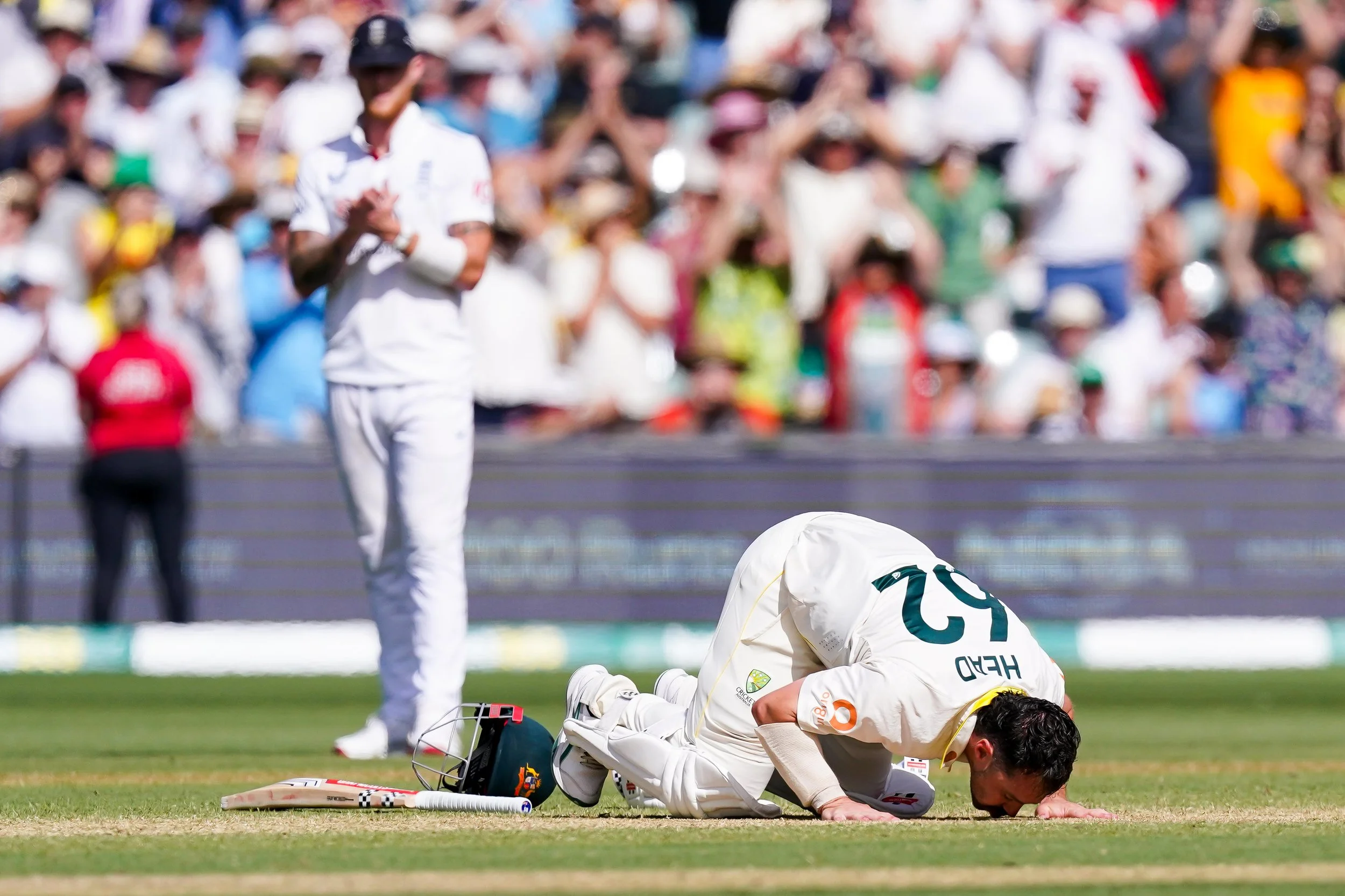 Cricket player kneeling on the ground with his head bowed and hands on the ground in front of him, another player standing nearby clapping, and a crowd of spectators in the background.