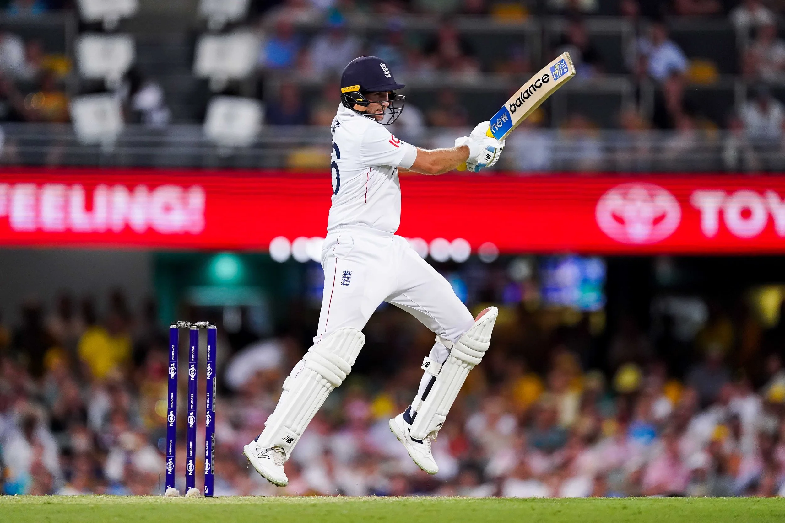 A cricket player in white uniform and helmet jumping in the air after hitting the ball with a bat, in a stadium with an audience.