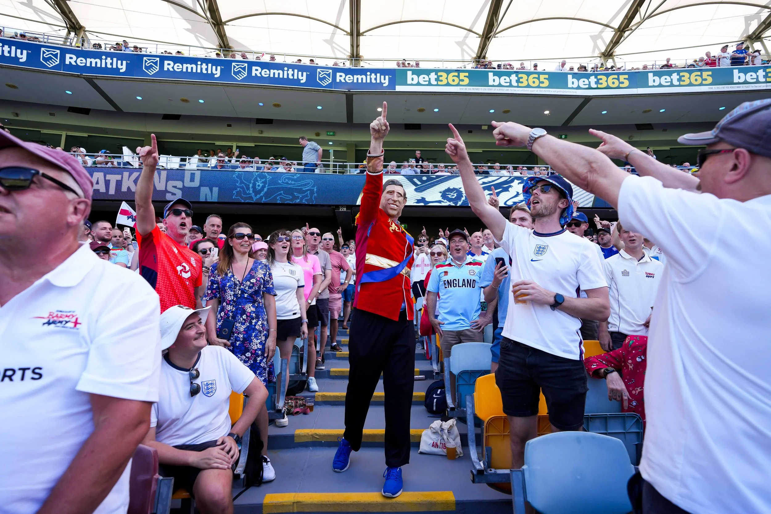 Crowd of cricket fans in stadium, some standing and cheering, wearing England team shirts, some pointing and taking photos, with banners and advertisements in the background.