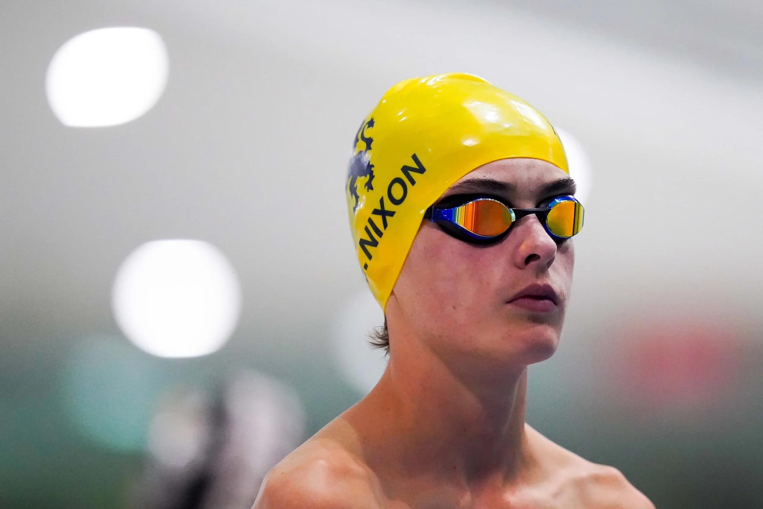 Young swimmer wearing a yellow cap with 'NIXON' branding and a lion logo, along with swim goggles with rainbow-colored reflective lenses, preparing for a swim in an indoor pool.