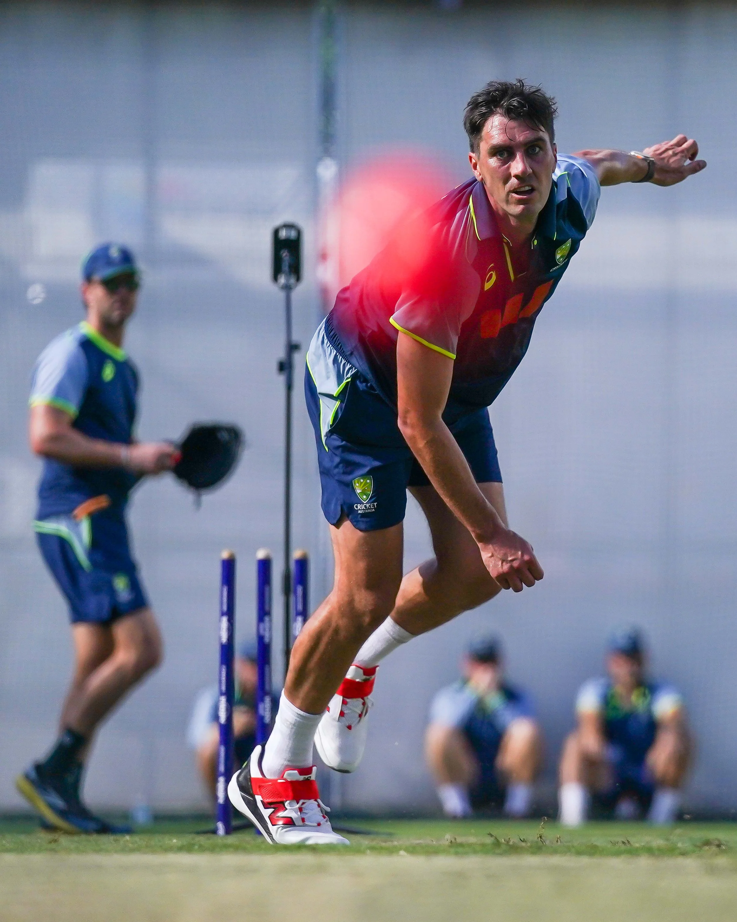 A male athlete in a blue sports uniform with yellow accents and white sneakers is bowling in a sports match, with a red glow near his face indicating intense focus, while another person in similar sportswear is observing in the background.