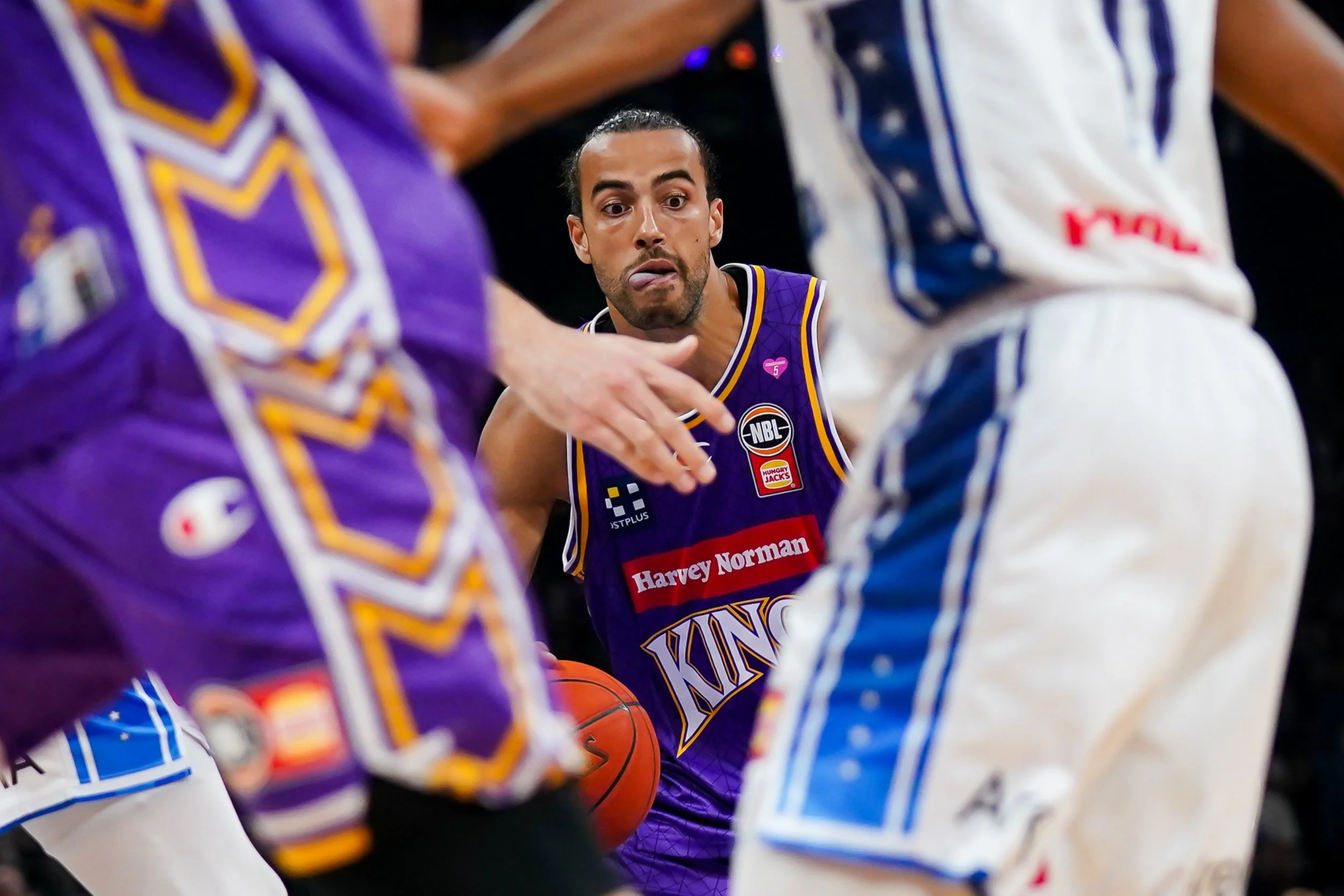 Basketball player in purple jersey holding ball surrounded by two opponents in white and purple jerseys during game