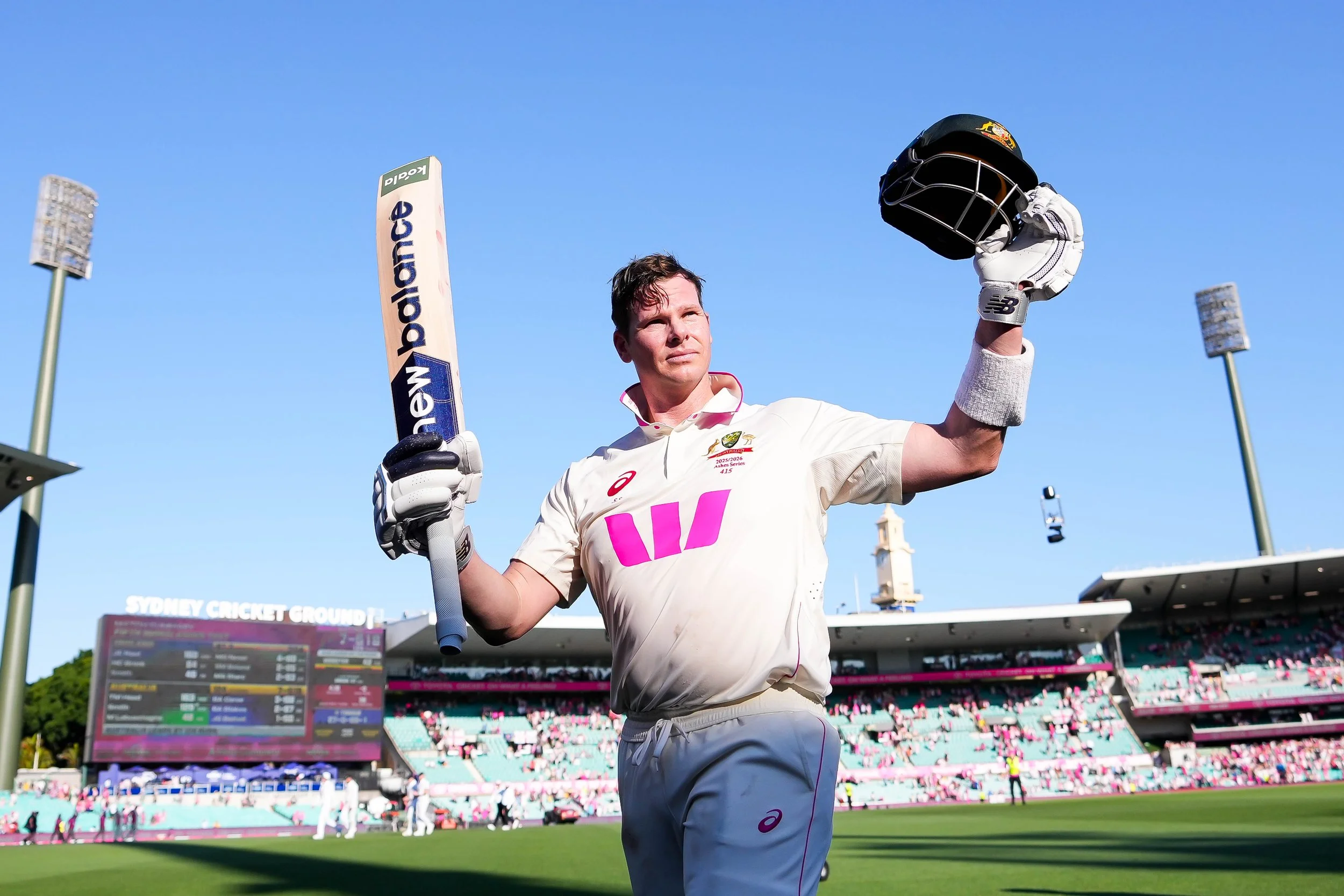 Cricketer standing on cricket field holding helmet and bat."