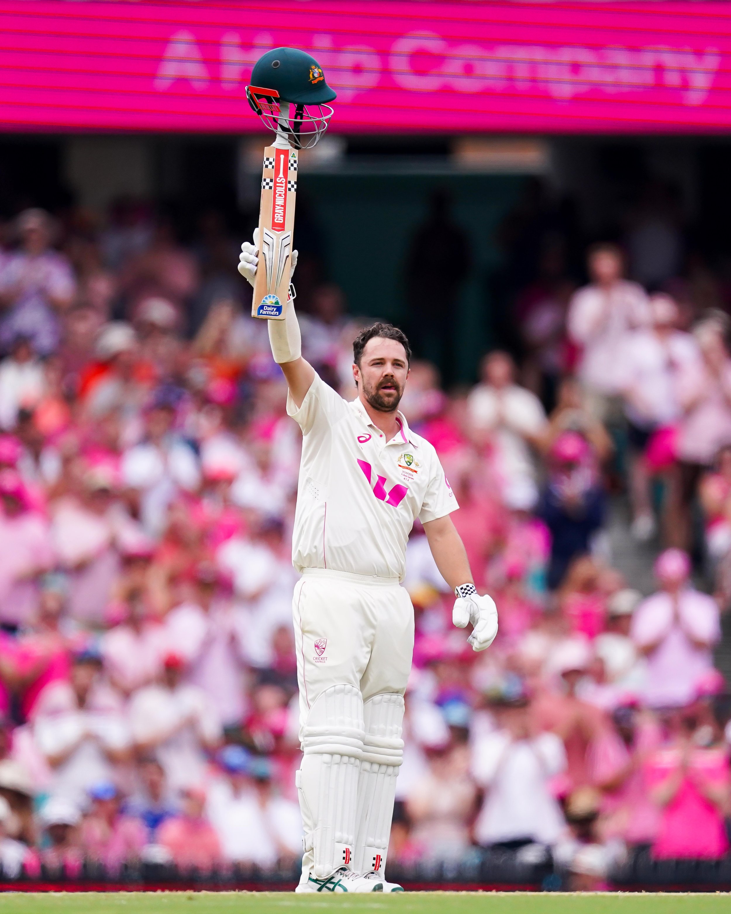 Cricketer celebrating by raising his bat and helmet during a match, with spectators in the background.