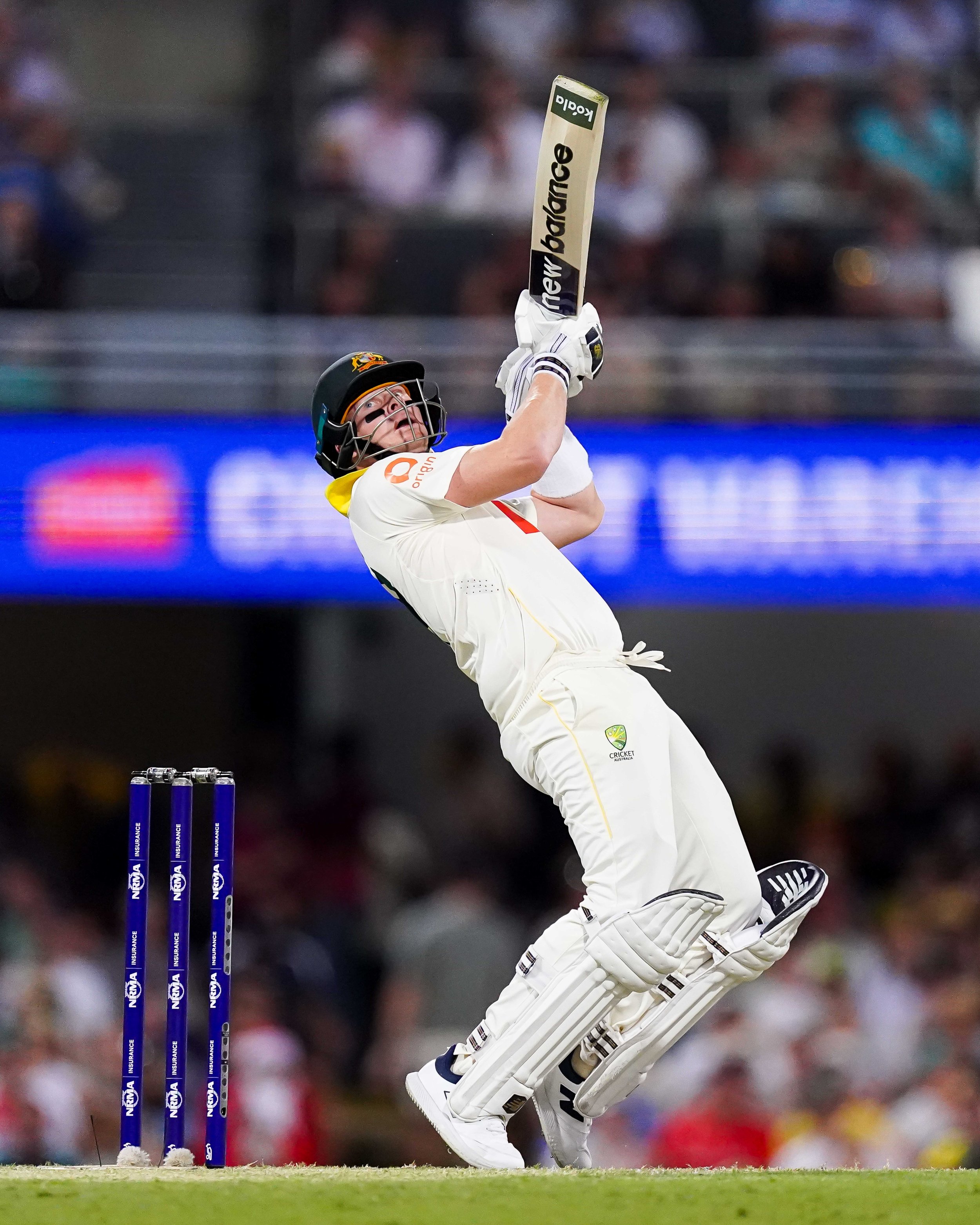 A cricket player in white uniform and helmet, holding a bat raised high, appears to have hit the ball successfully during a match.