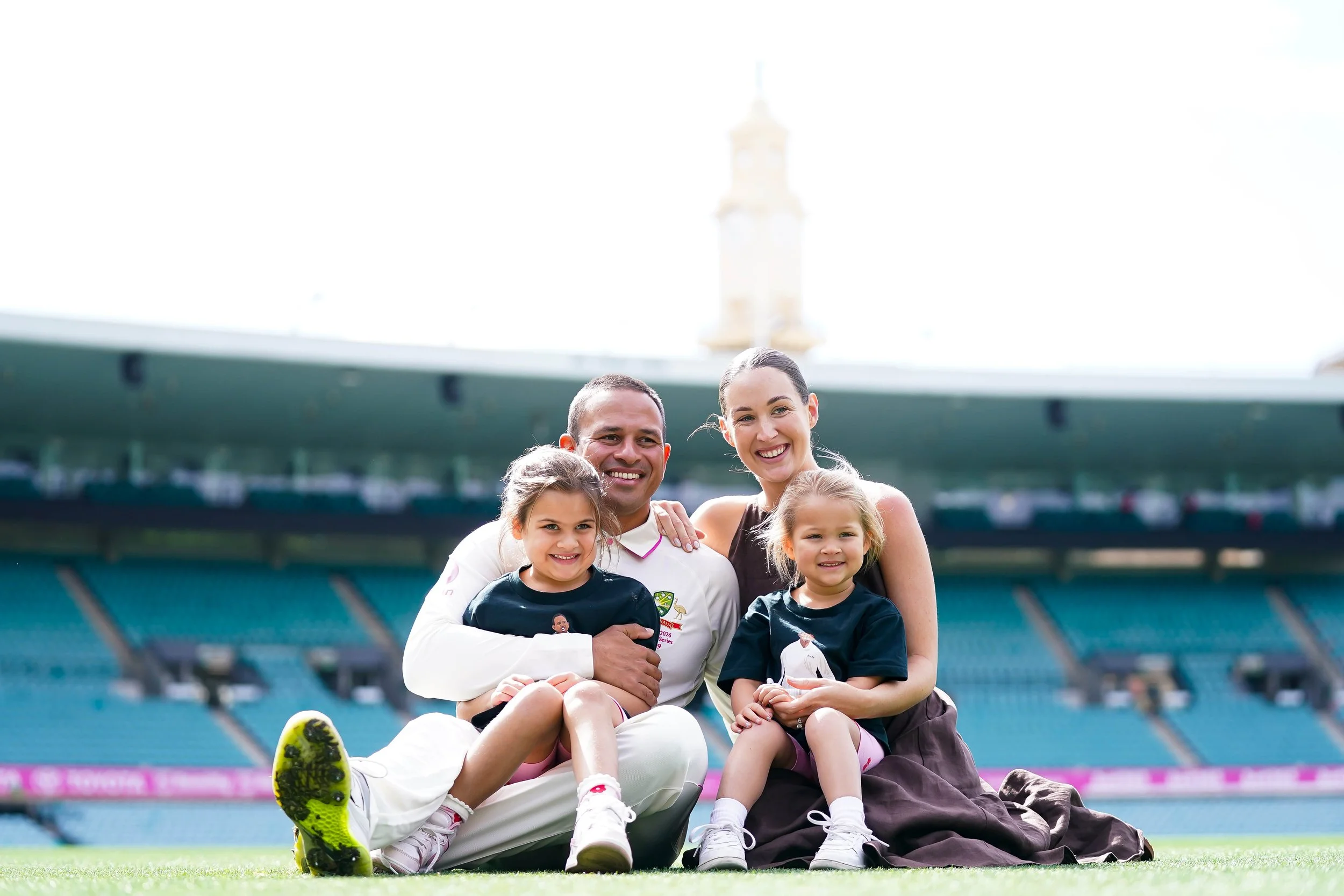 Family sitting on the field of a sports stadium, celebrating. The family includes two women, a man, and two young girls, all smiling and posing together.