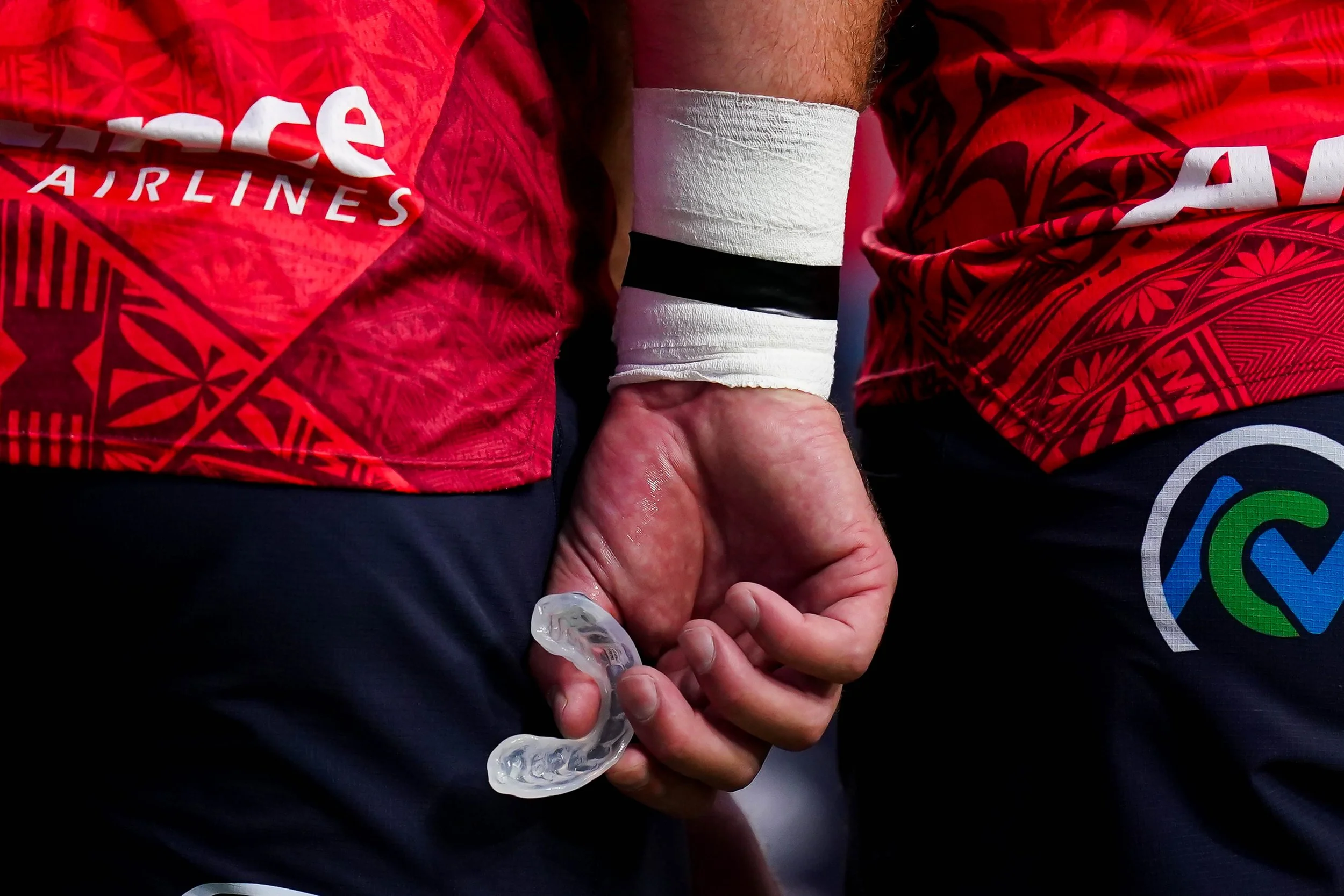 Close-up of a rugby player's hand holding a clear mouthguard, with other players in red jerseys in the background.