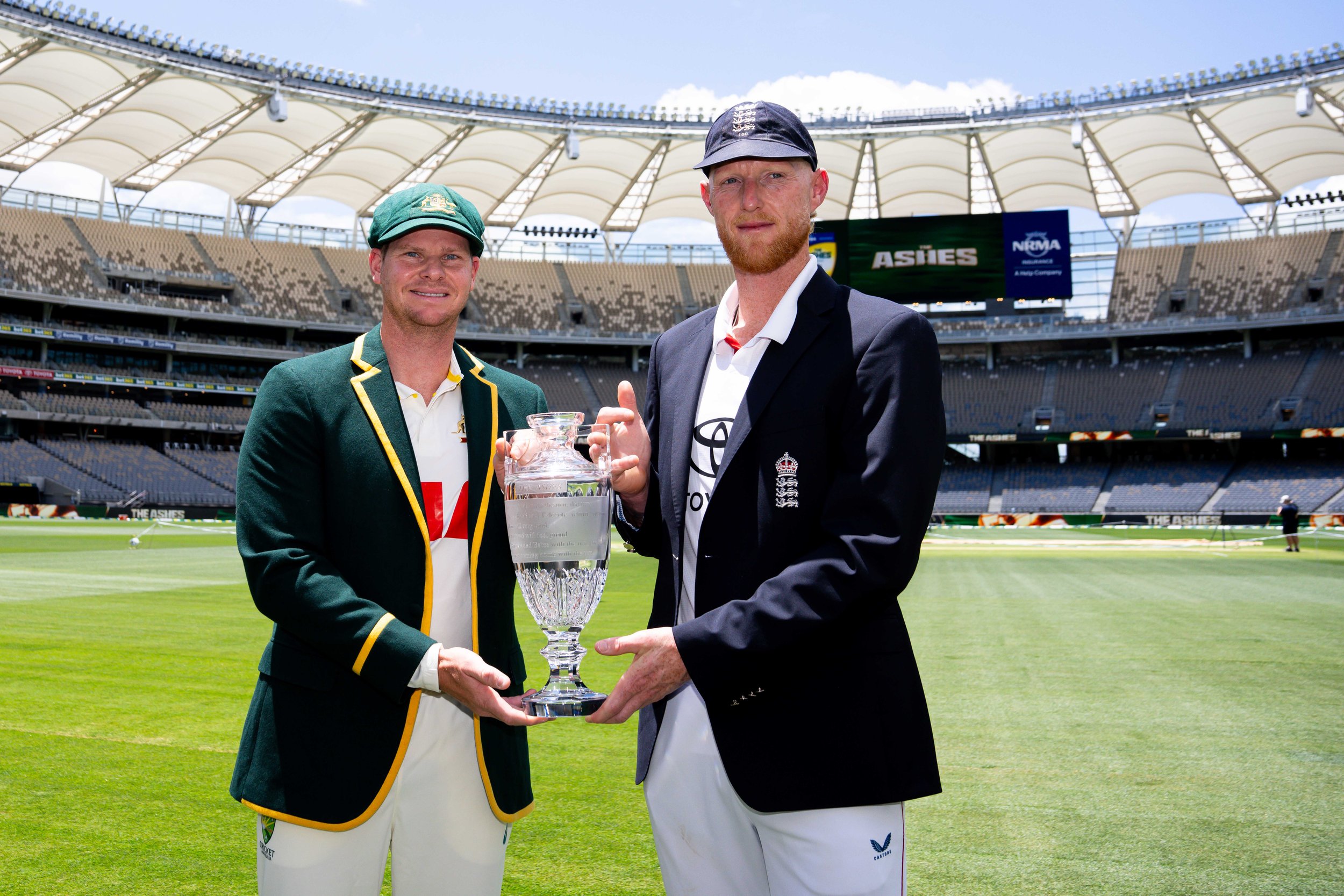 Two cricket players in uniform holding a large trophy on the field of a stadium.