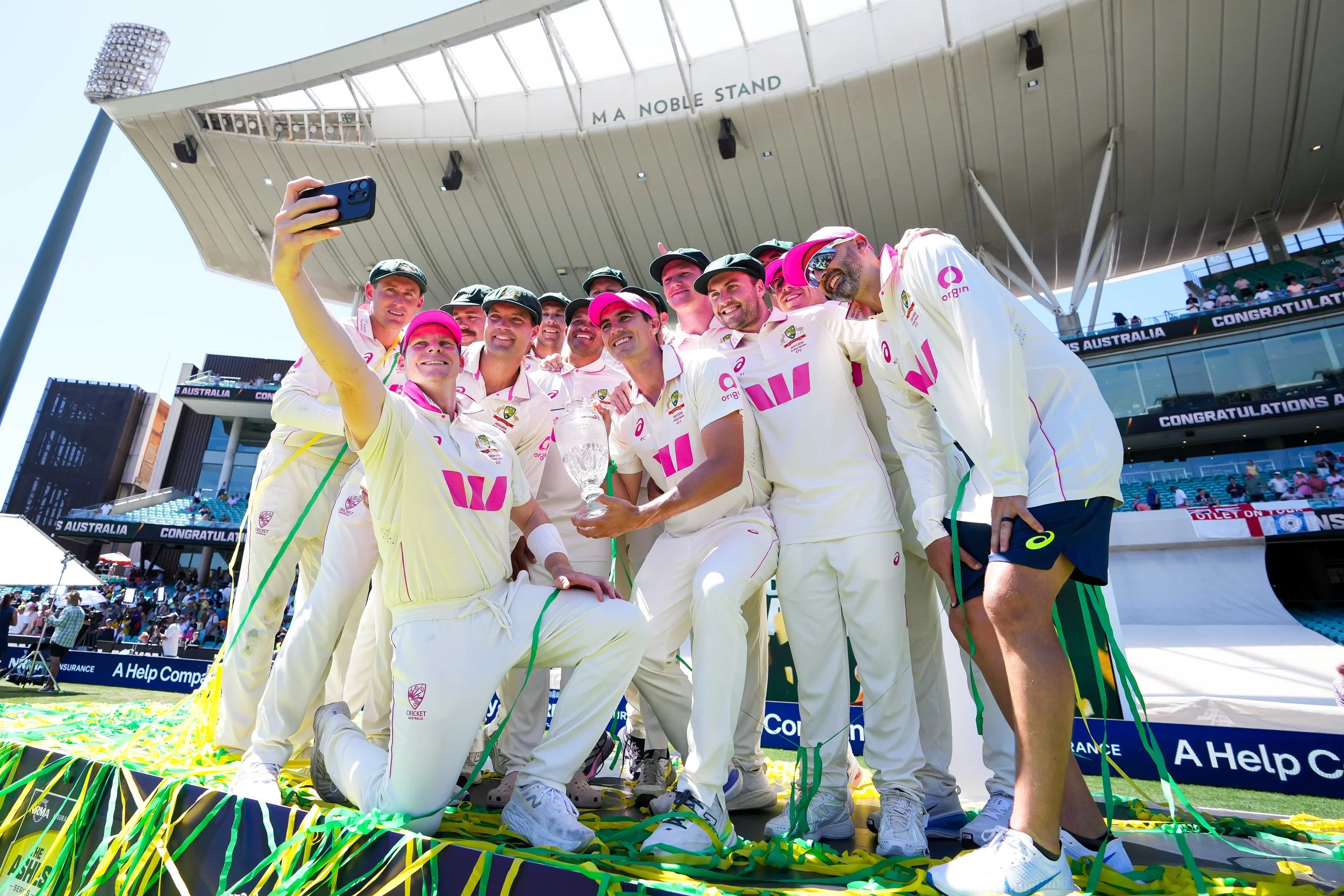 Australian cricket team celebrating with a trophy on a cricket field, taking a selfie after winning a match.