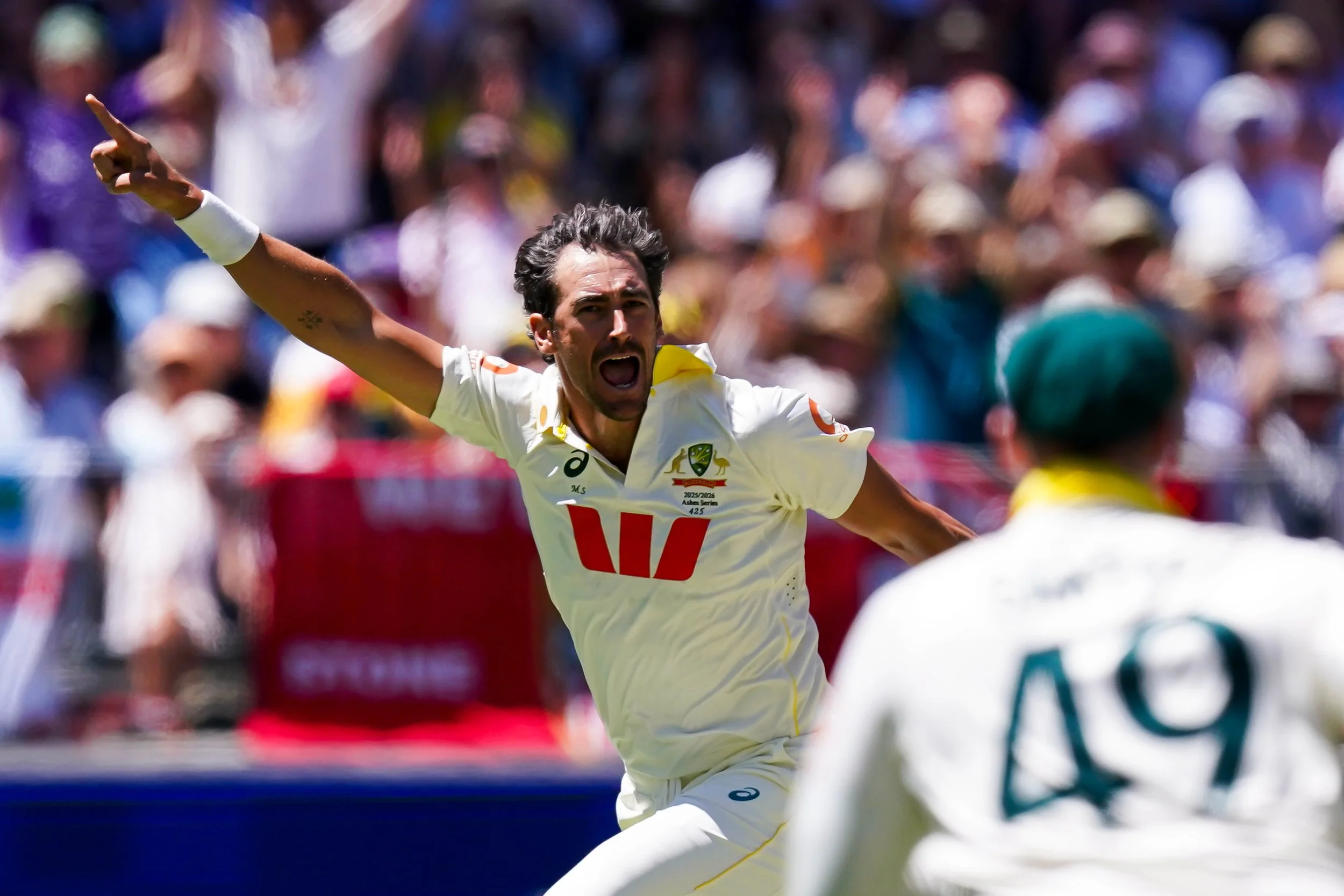 Mitchell Starc celebrating on the field with a passionate expression, wearing a white uniform with the Australian national team logo, with a crowd in the background.