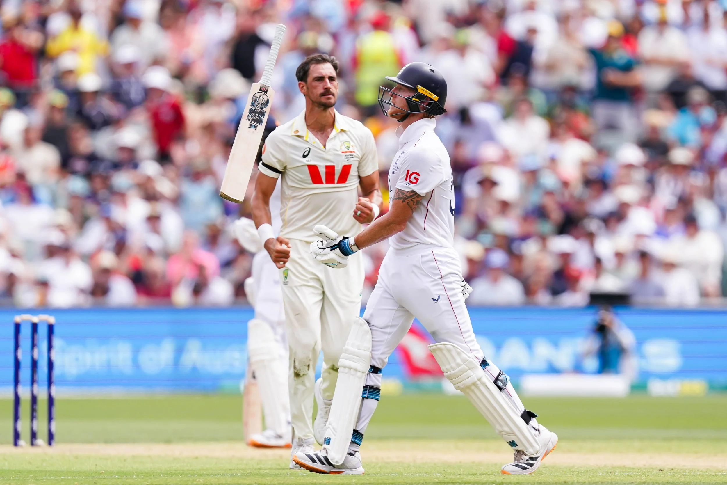 Two cricket players on the field during a match, one wearing a helmet and batting gear, the other in a white uniform, with a crowd in the background.