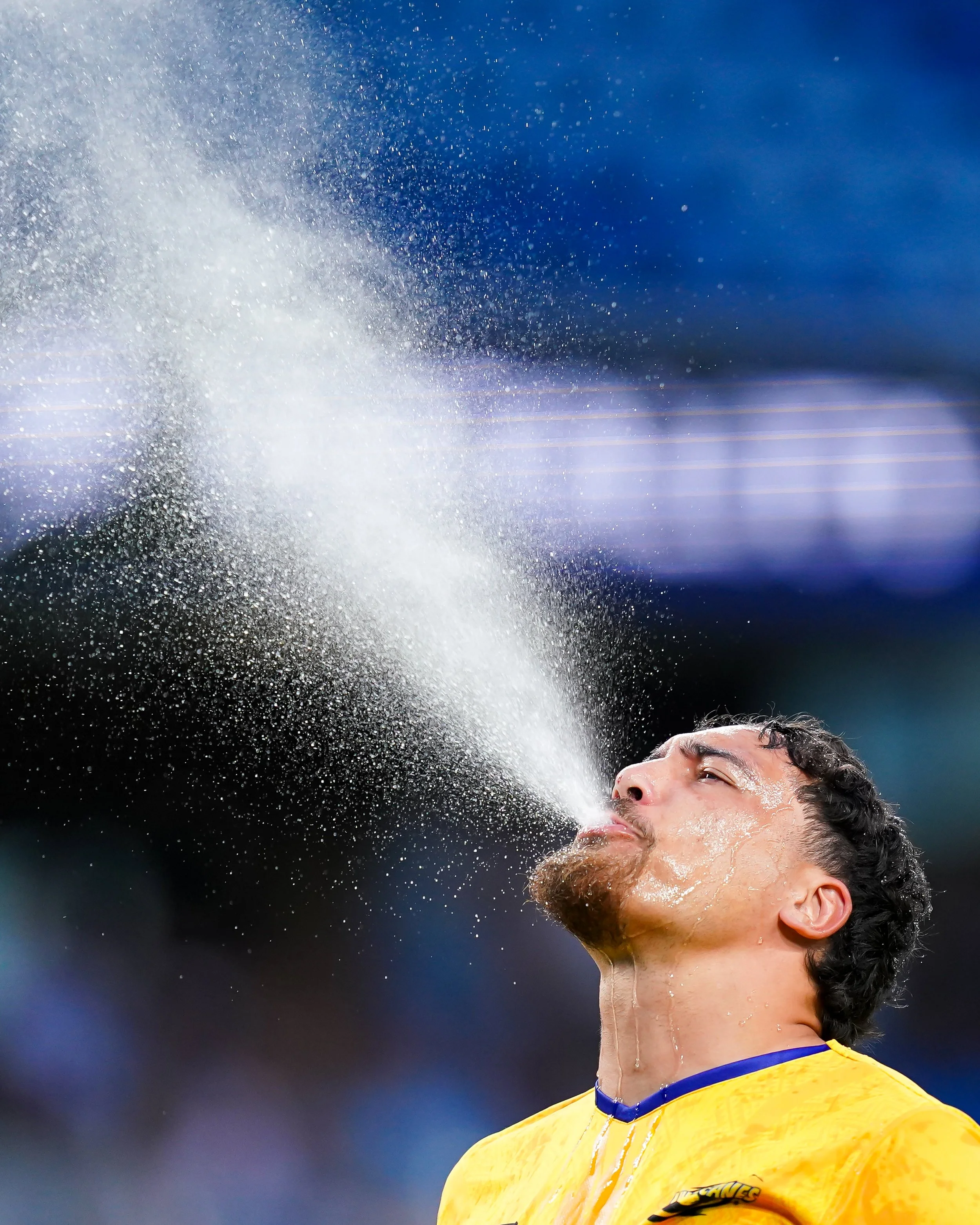 Soccer player in yellow jersey spitting water during a game with a blurred stadium background.