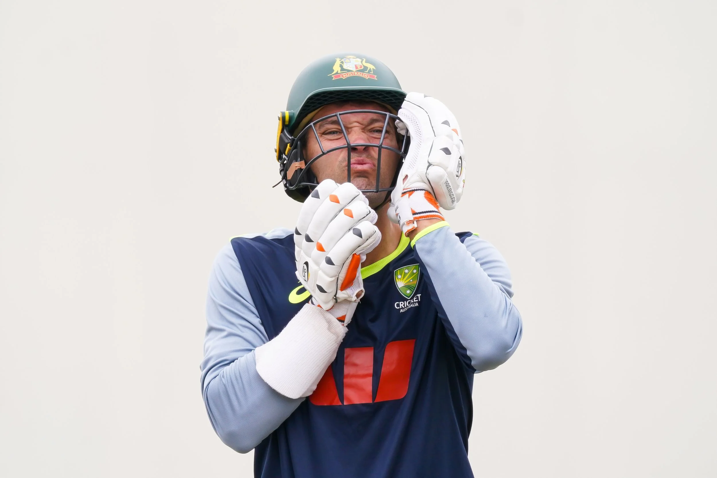 A cricket player wearing a helmet and gloves, adjusting his helmet during practice or game, with the Australia national cricket team logo visible on his jersey.