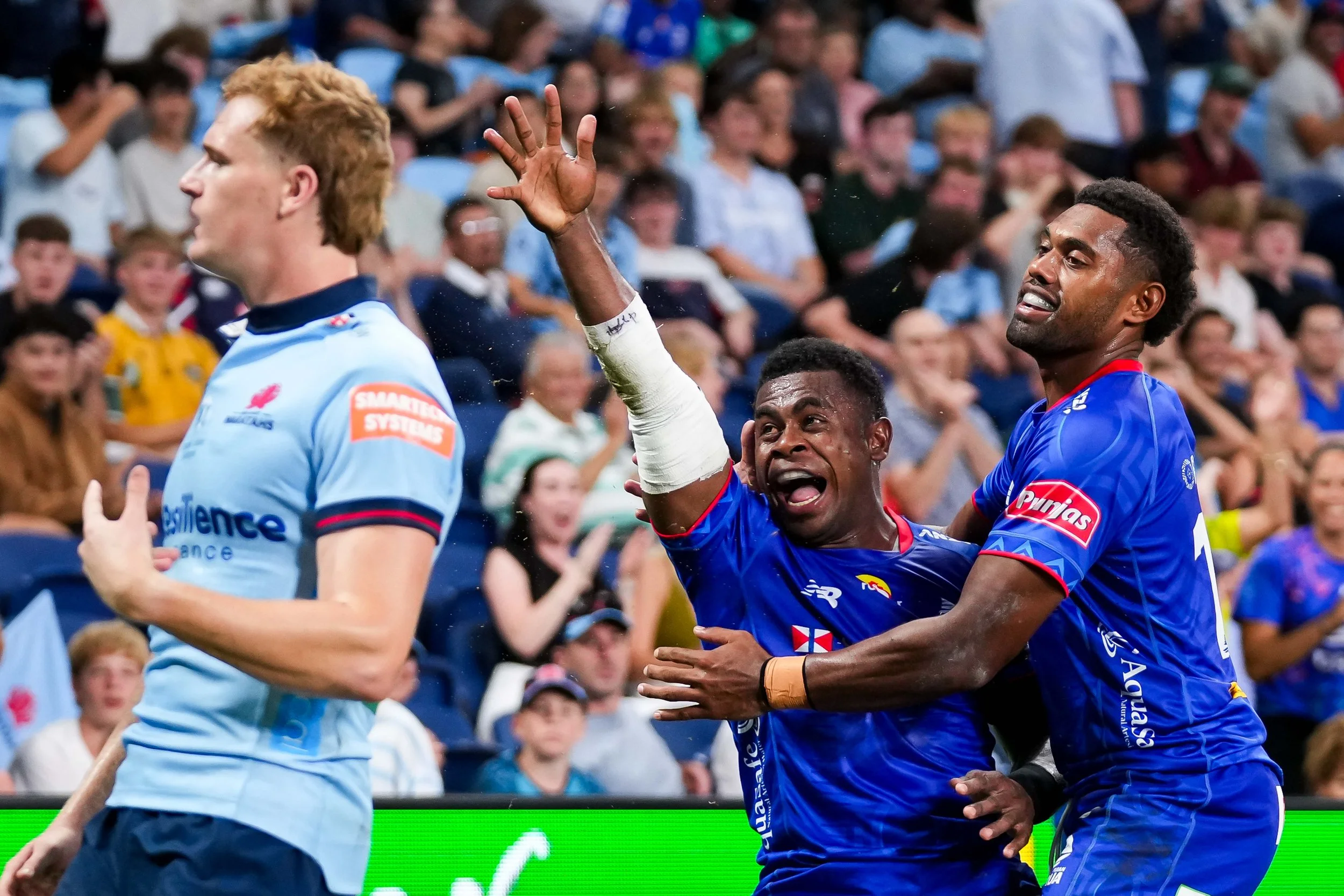 Cricket players celebrating a successful play on the field with a crowd in the background.