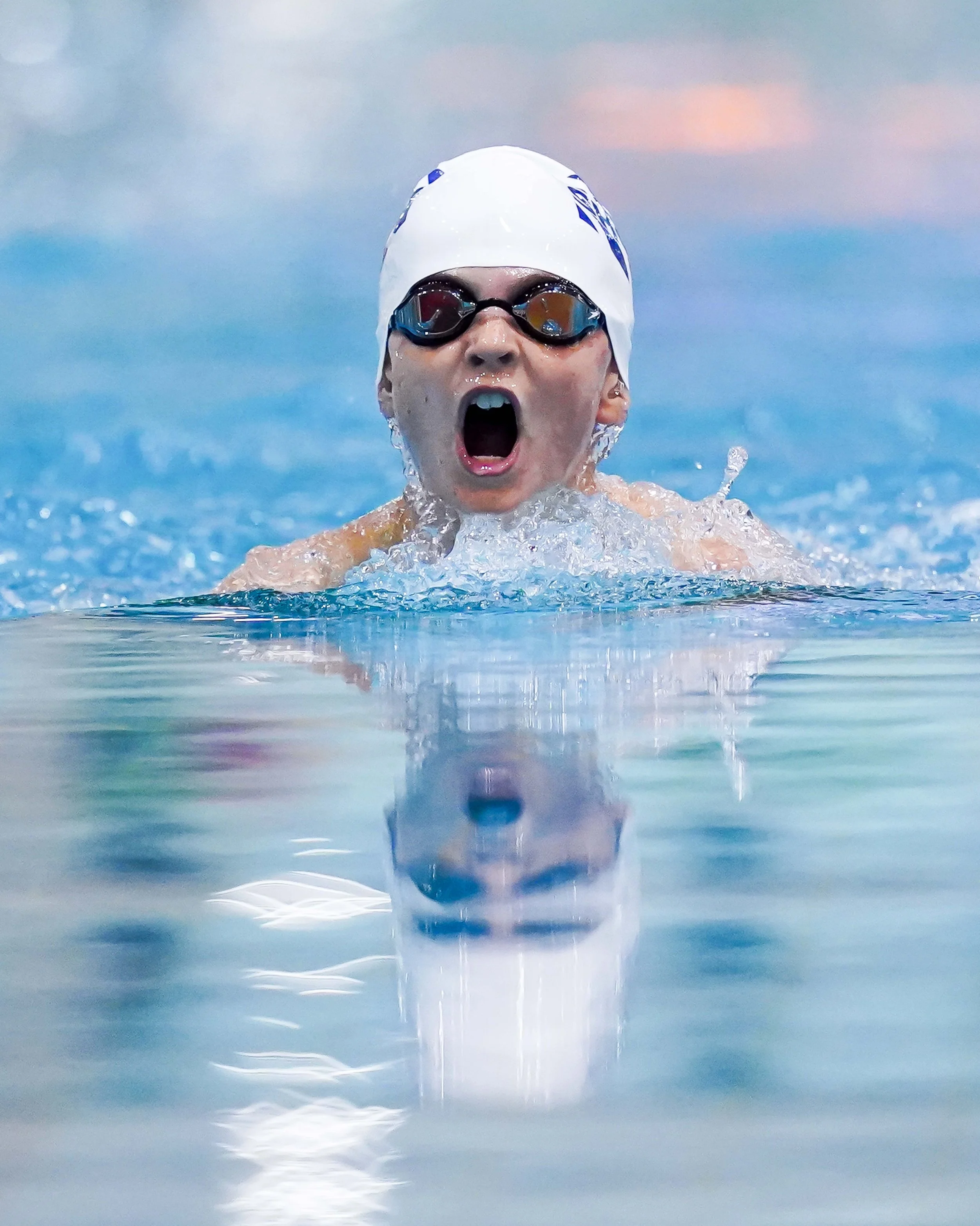 A young swimmer in a white swim cap and goggles is swimming in a pool with their mouth open, possibly shouting or breathing heavily, with their reflection visible in the water.
