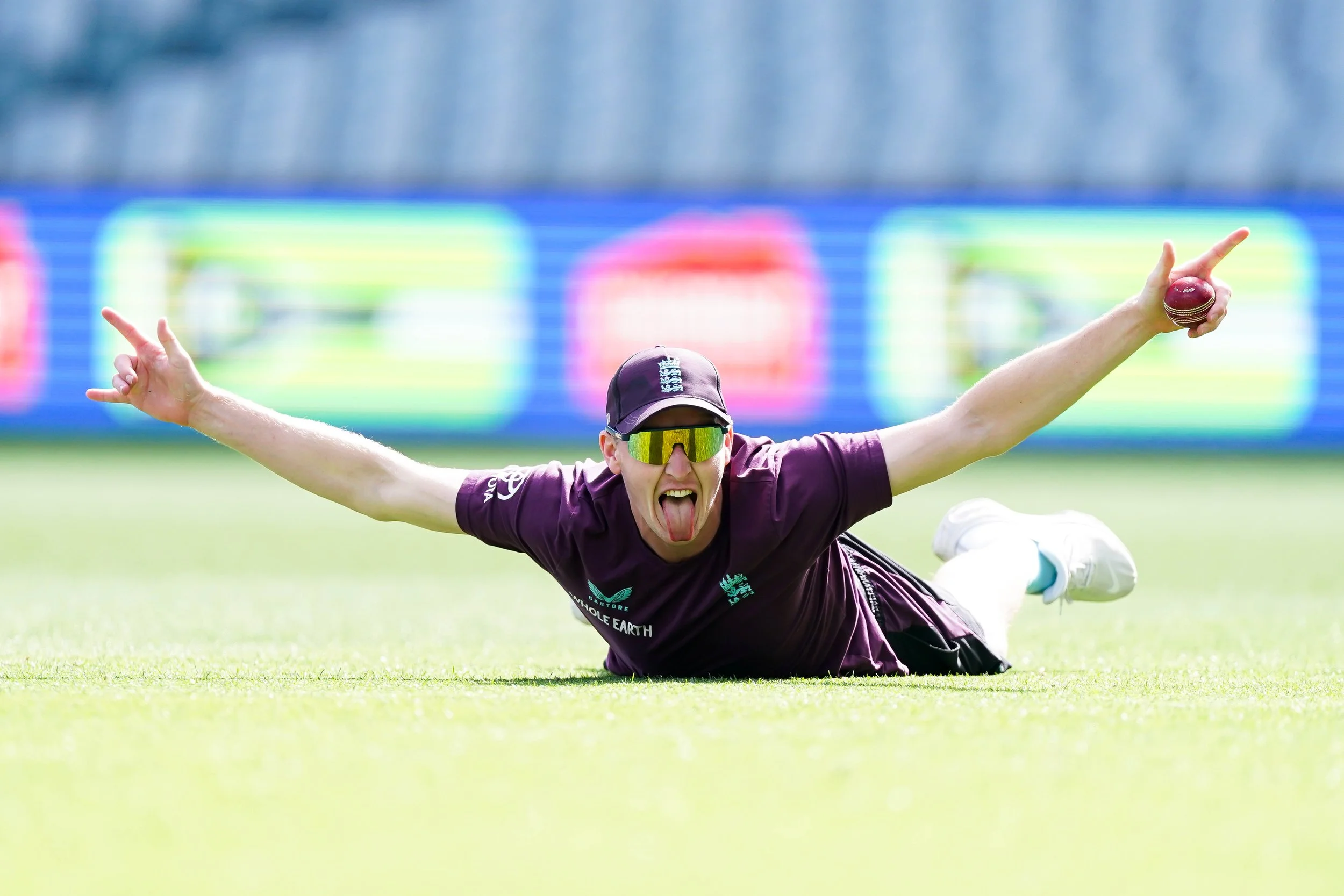 Cricket player in maroon uniform diving on the field with arms outstretched, holding a cricket ball, tongue sticking out, and wearing sunglasses during a game.