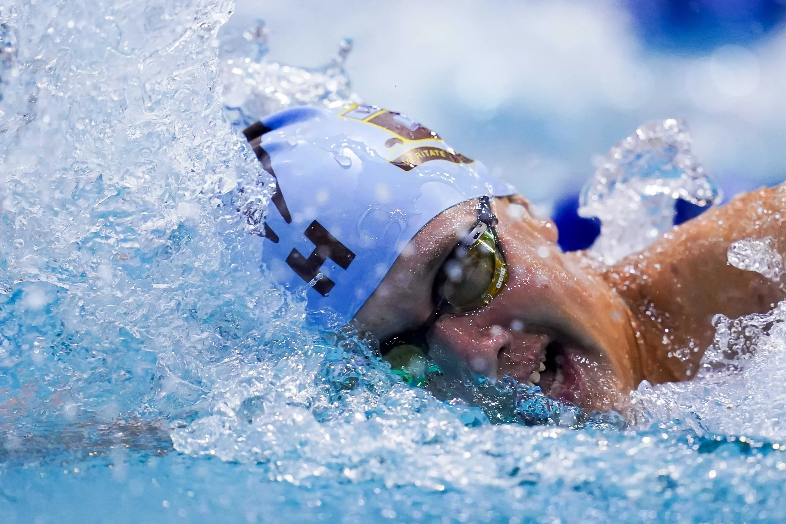 A swimmer wearing a white cap with black letters and goggles is swimming in a pool, surrounded by splashing water.