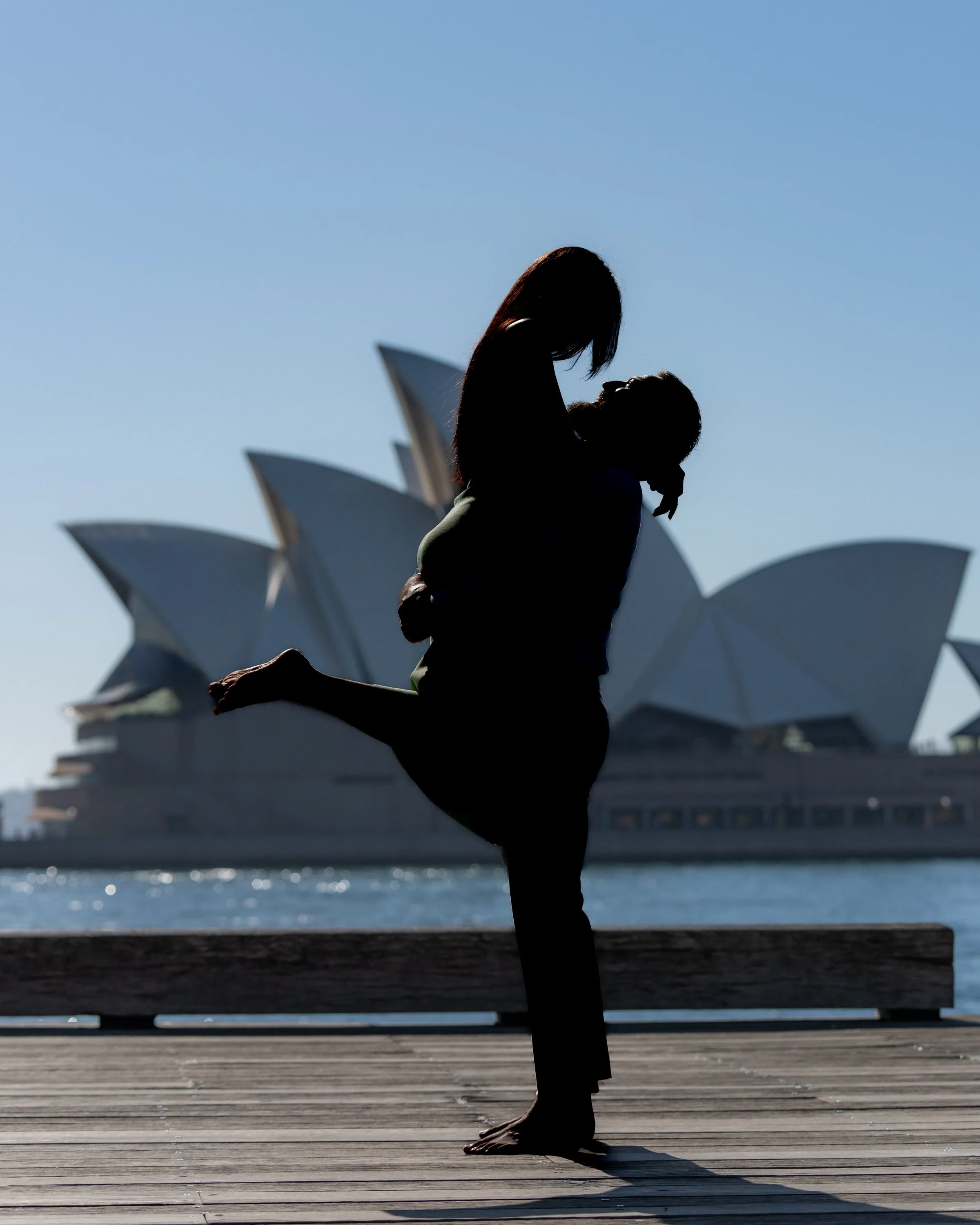 Silhouette of a woman lifting a child in front of the Sydney Opera House against a bright blue sky.
