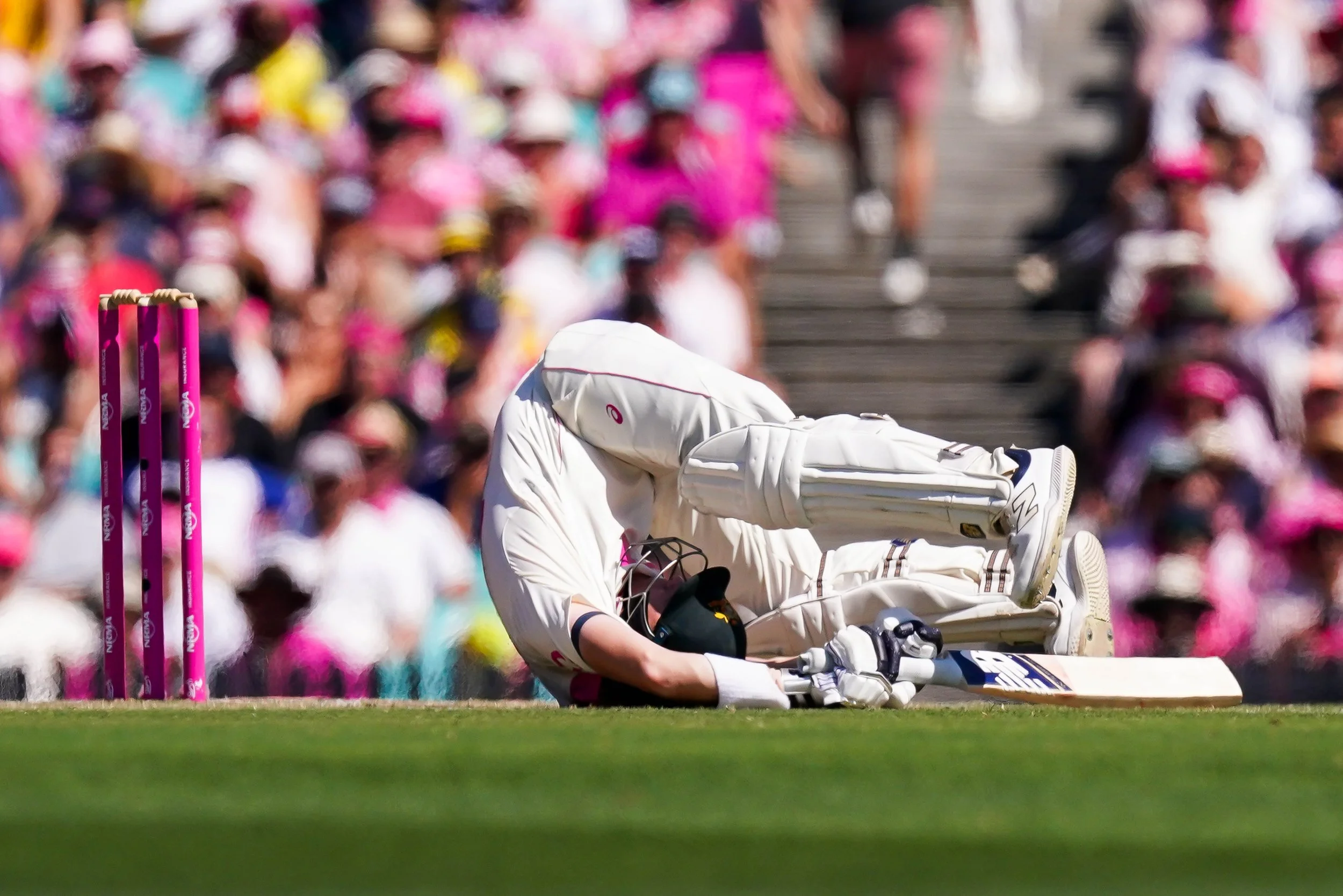 A cricket player in white uniform falling on the ground on a cricket field, with pink wickets nearby, during a match with spectators in the background.
