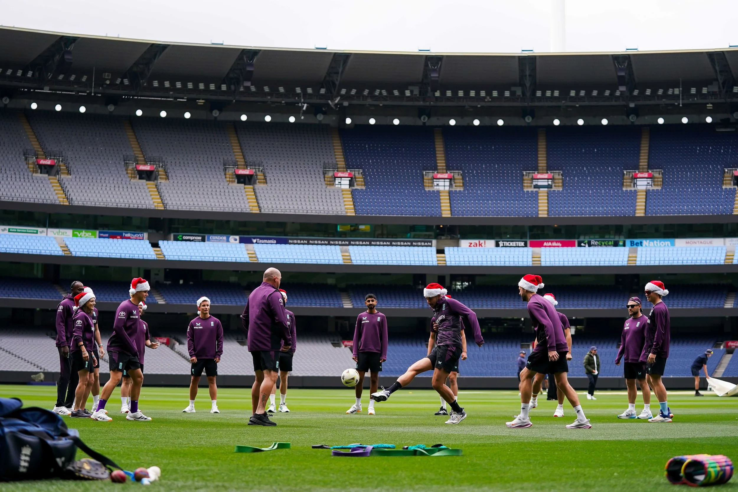 Cricket players wearing purple training gear and Santa hats practicing on a field in a large stadium, The MCG in Melbourne.