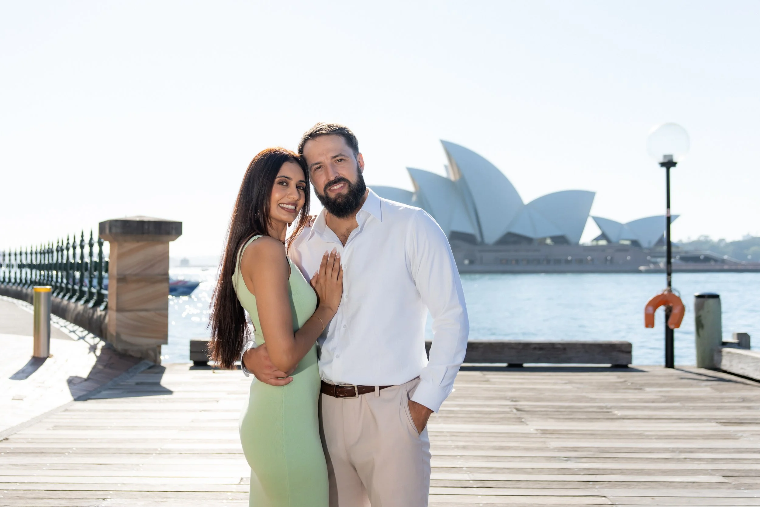 A smiling couple standing on a wooden dock near the Sydney Opera House, with the woman in a green dress and the man in a white shirt and beige pants, embracing each other.