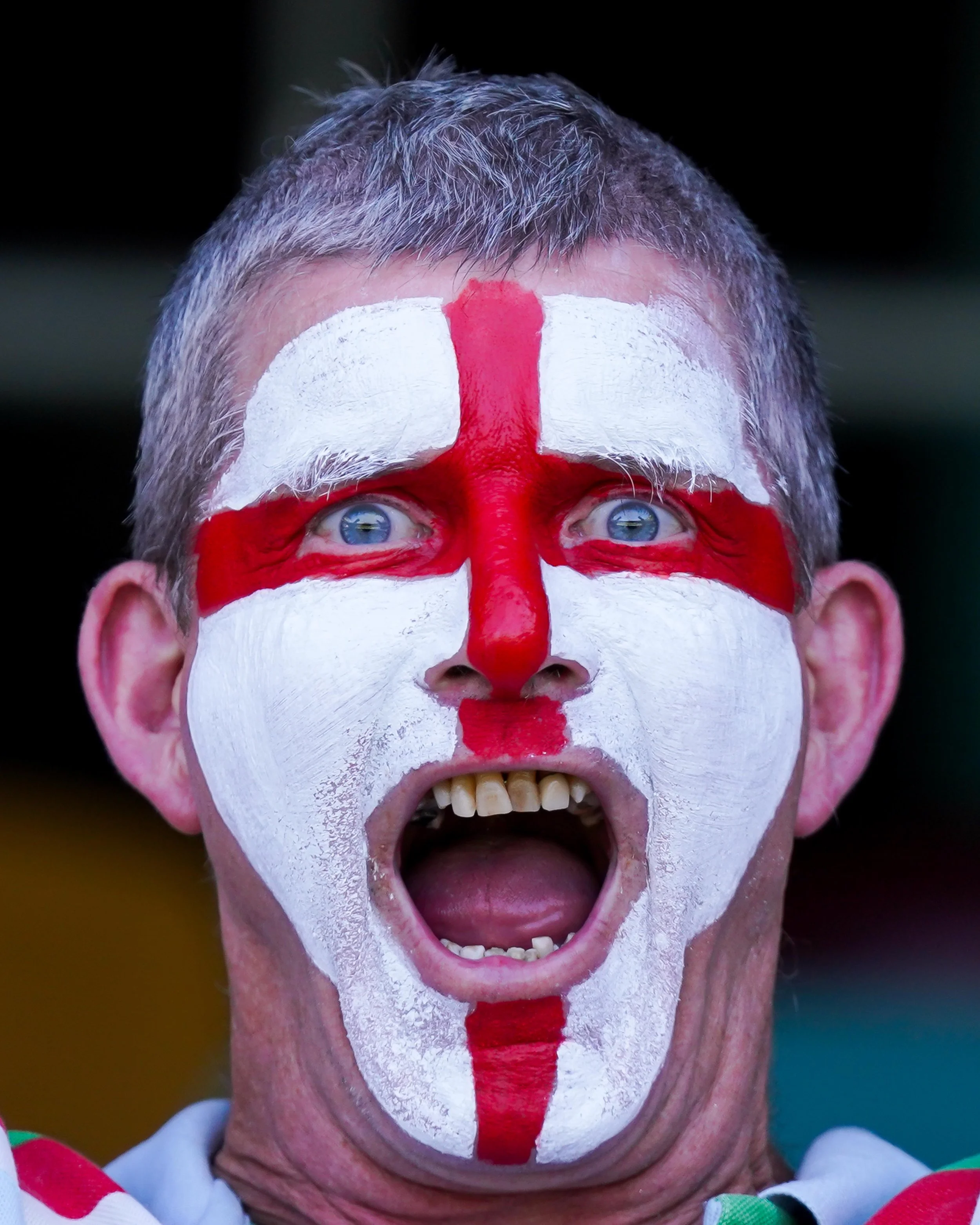 A man with face paint resembling the English flag, celebrating passionately with his mouth open.