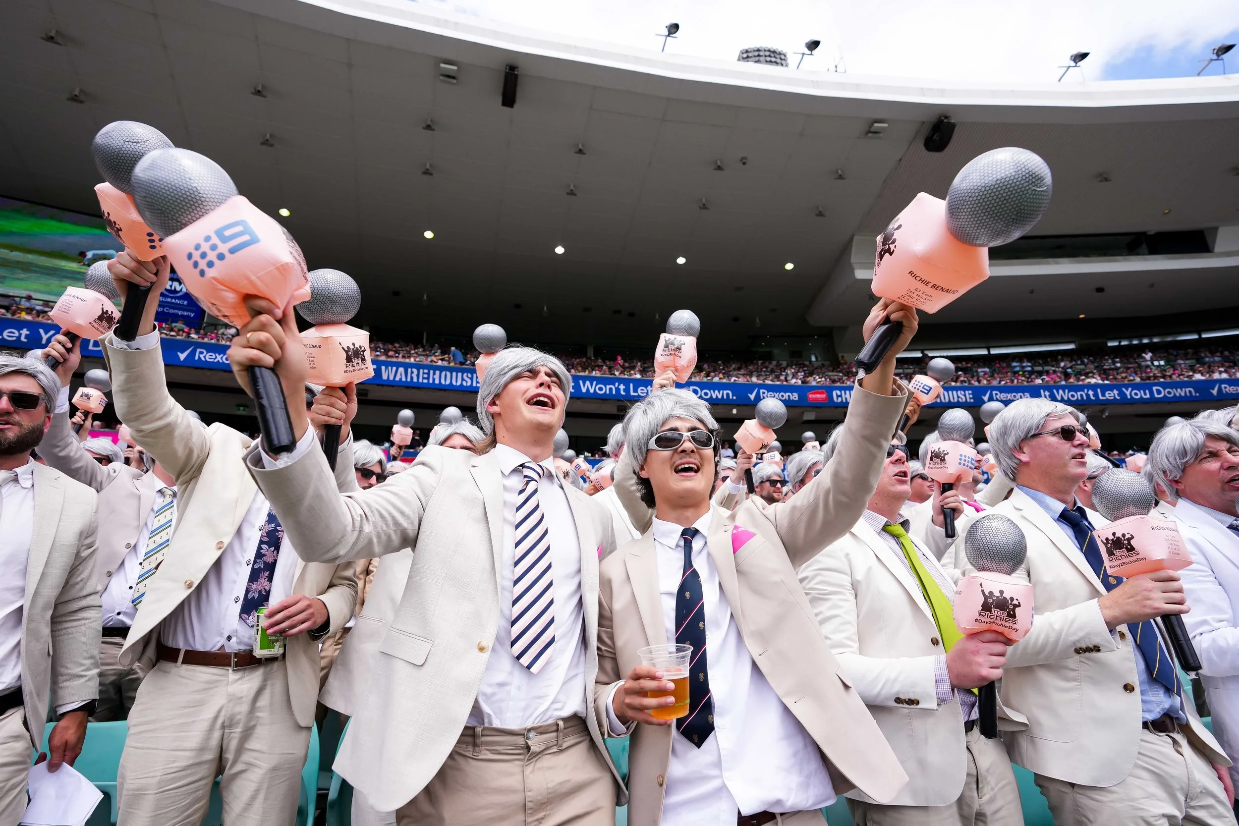 People dressed in beige suits celebrating at a stadium, holding microphones with pink foam covers, cheering and enjoying an event.