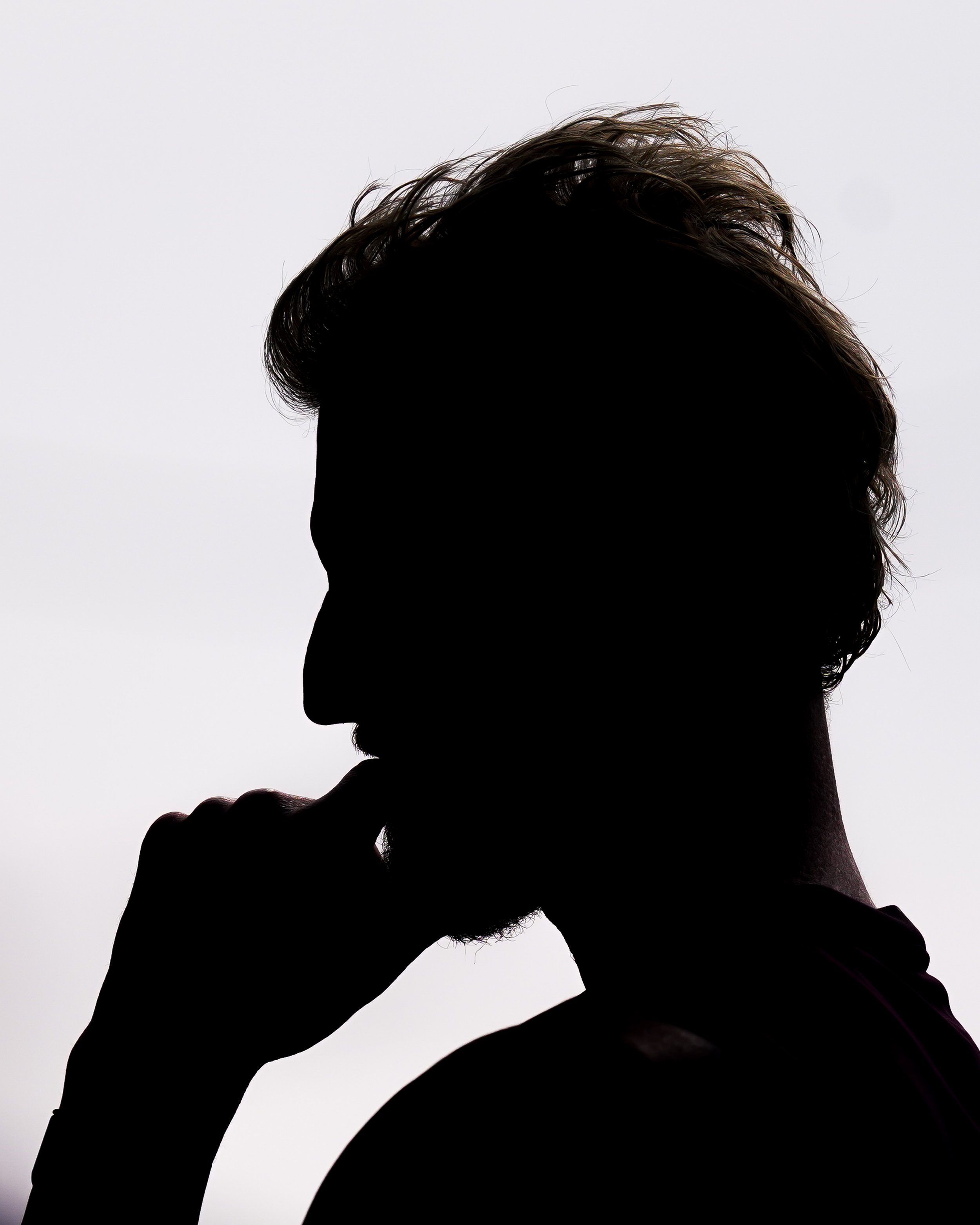 Silhouette of a man, Ben Stokes, with short hair and a beard, posed in profile with his finger touching his lips over a light background.